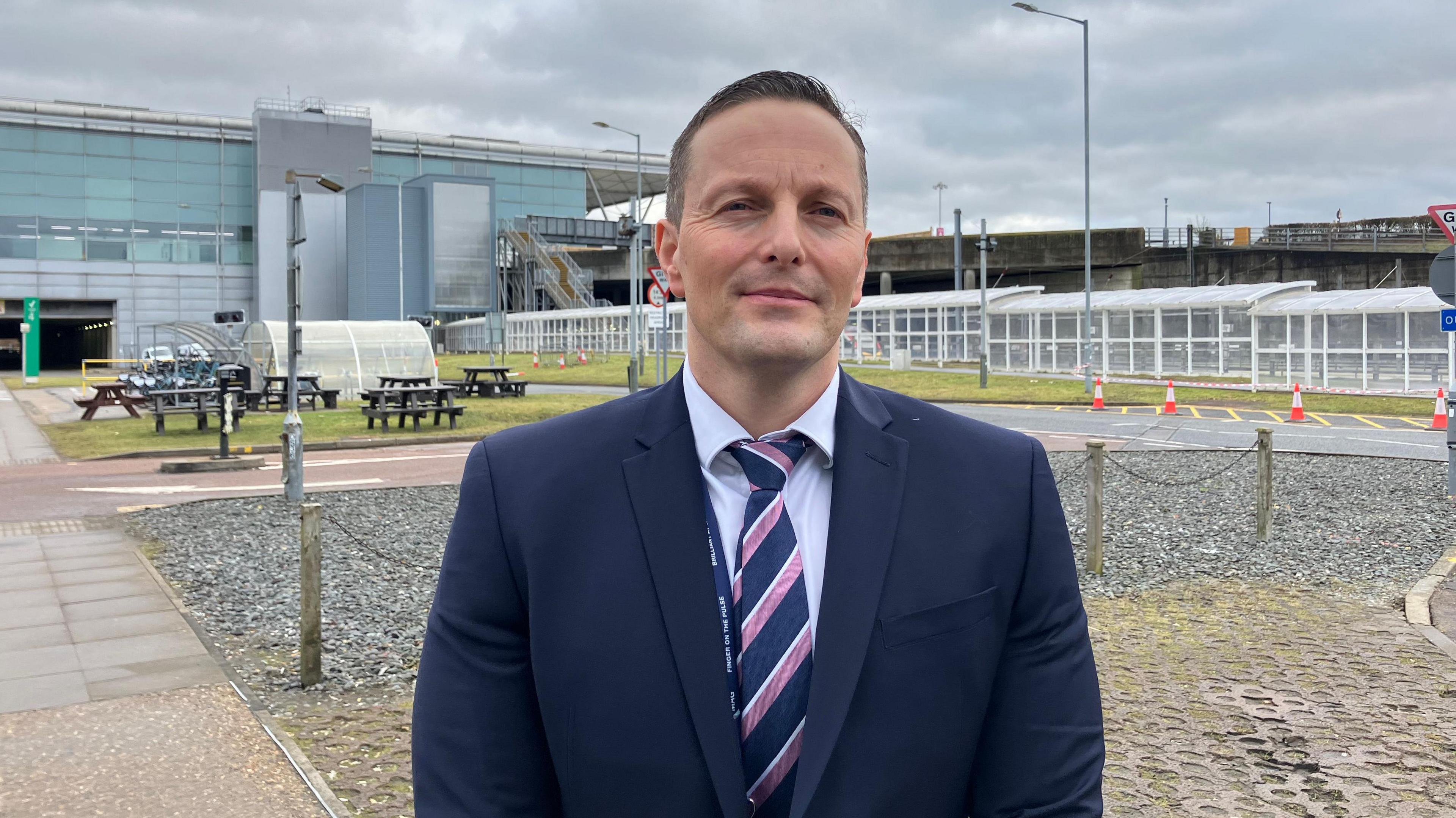 A head and shoulders picture of Alex Adams wearing a suit and tie outside Stansted Airport