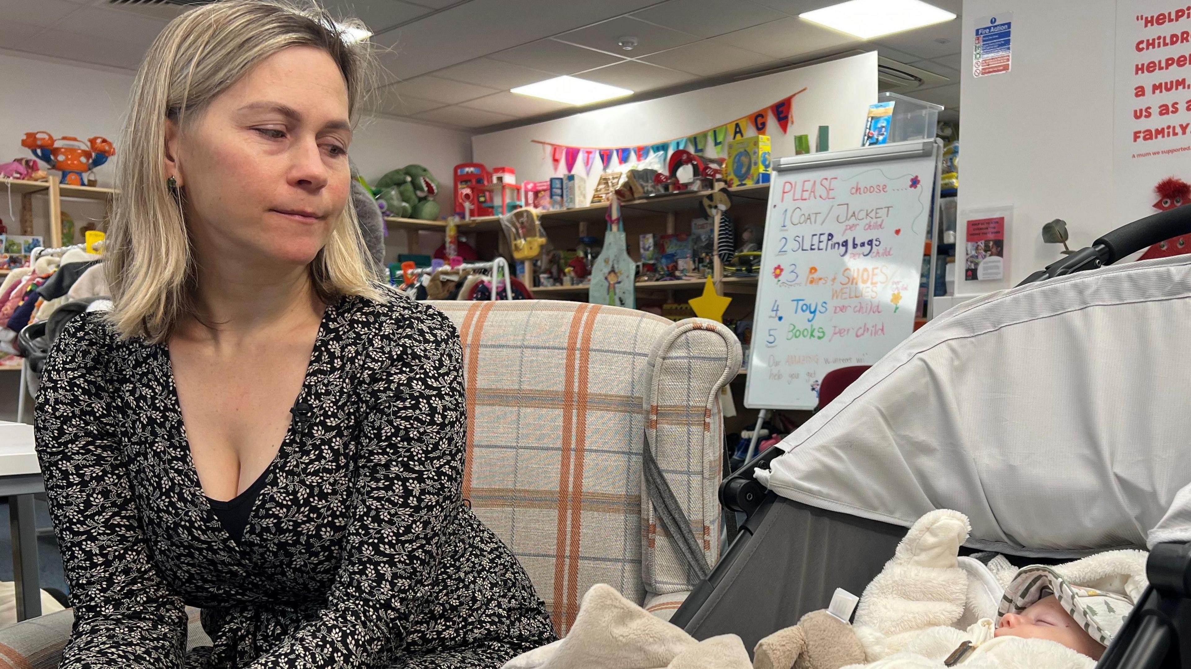 Nataliia Kravchenko, wearing a long-sleeved black top with small flowers on it, looks into the pram of her new born baby. Shelves of toys are in the background.