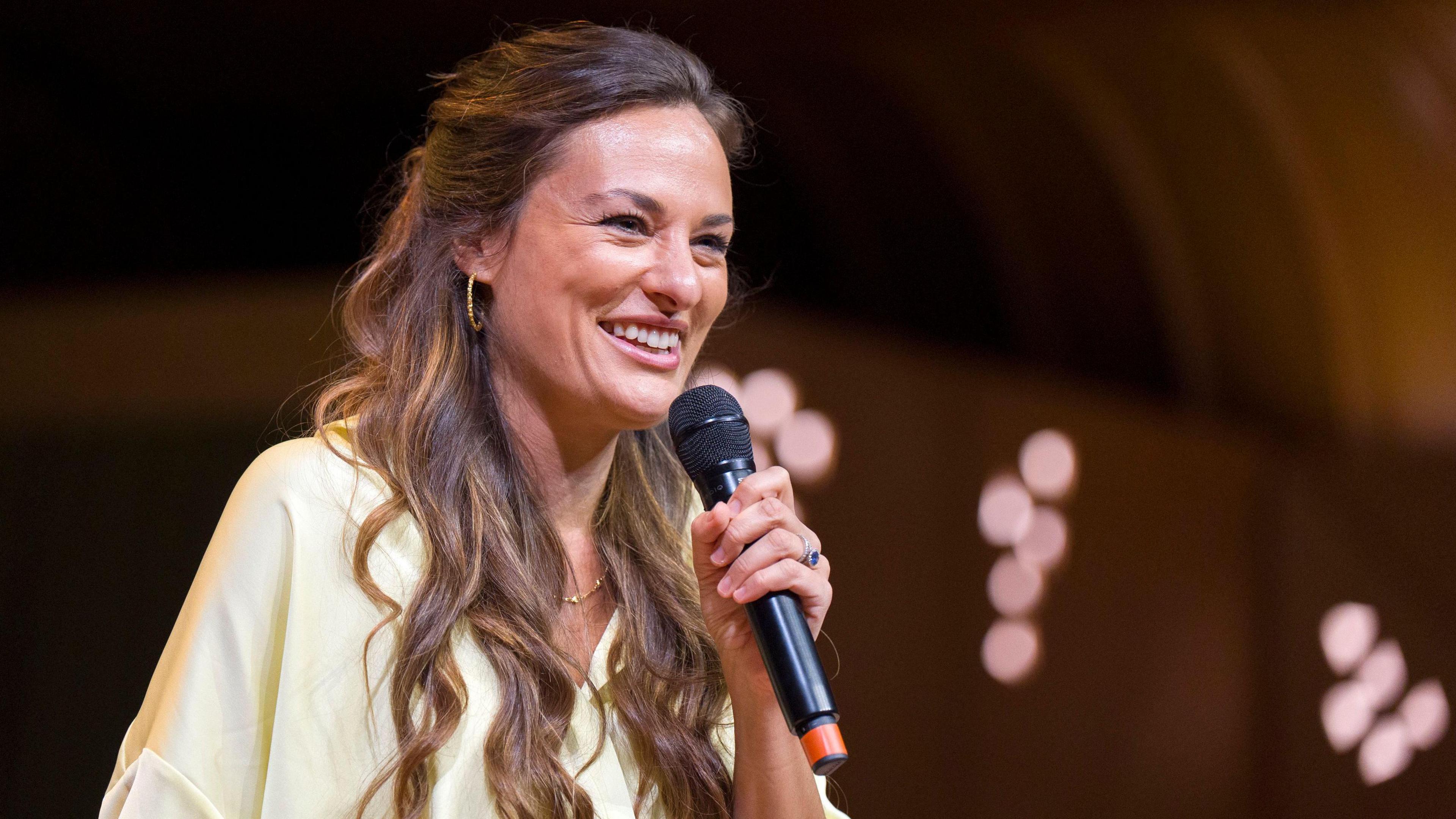 Nicola Benedetti smiling with dark hair. She is holding a microphone to her mouth with her left hand and is wearing a light yellow top.