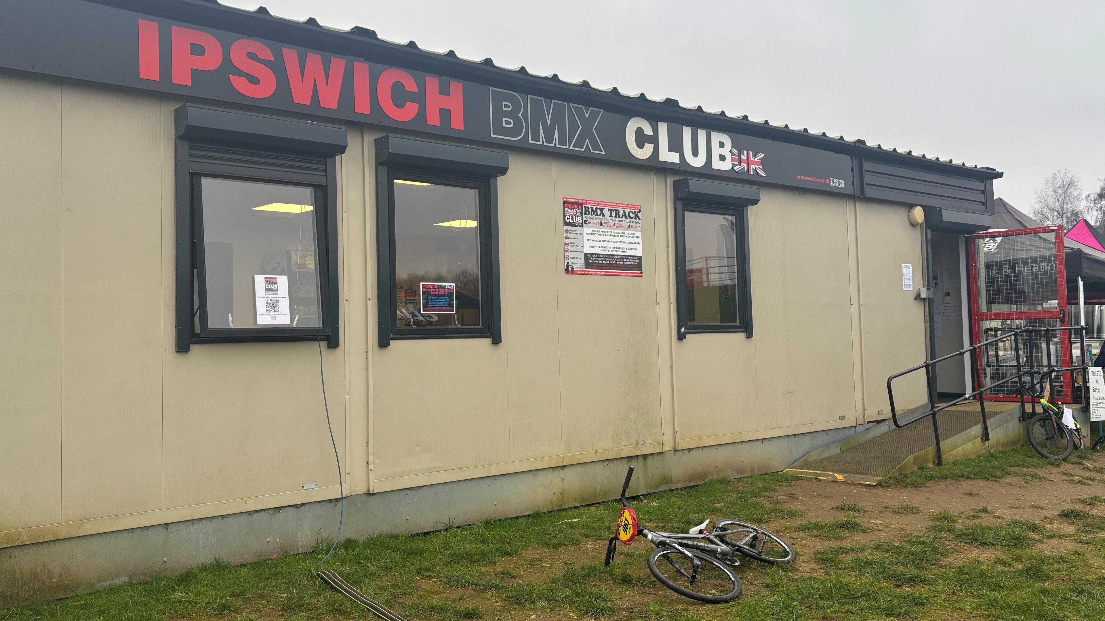 A pale clubhouse building with a black and red "Ipswich BMX club" banner across the top. A BMX bike with a red front lays in front of the clubhouse.