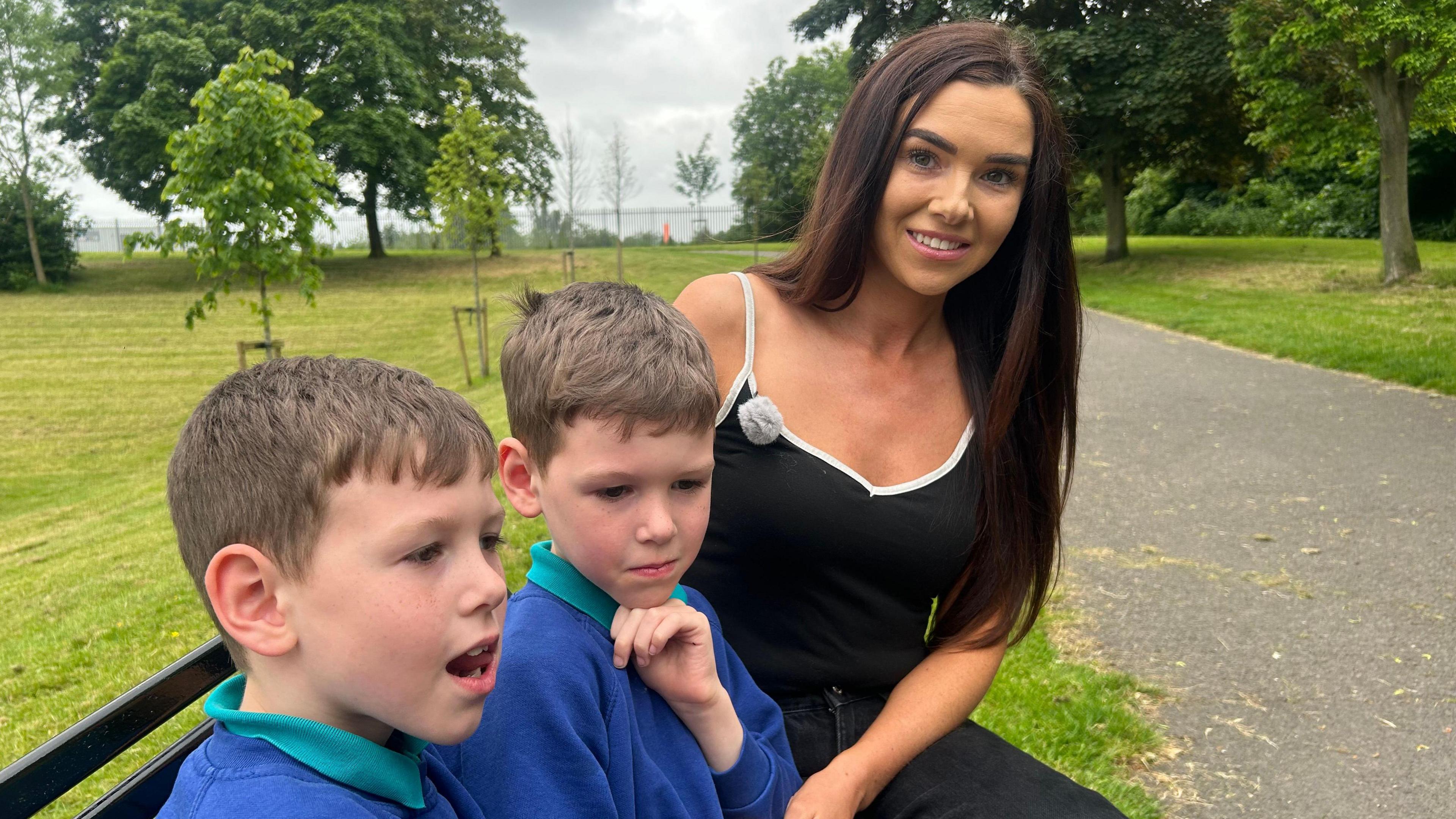 Megan is sitting on a bench outside with her two sons. She is wearing a black top, her sons are wearing a blue school uniform. There are trees in the background.