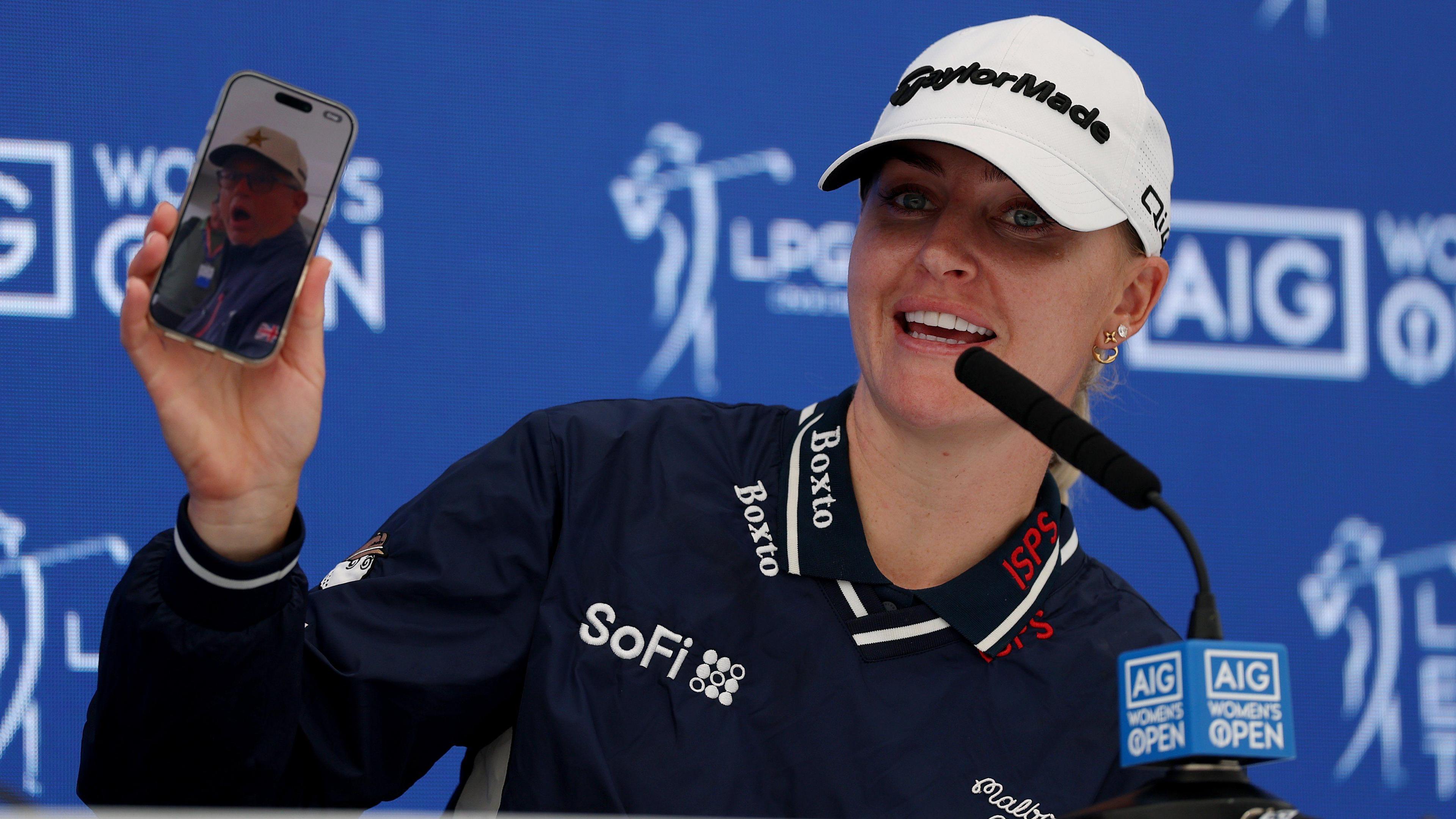 Charley Hull of England shows a video of her caddie Adam Woodward on her phone as she speaks to the media, explaining how she pranked him with a fake scratch card