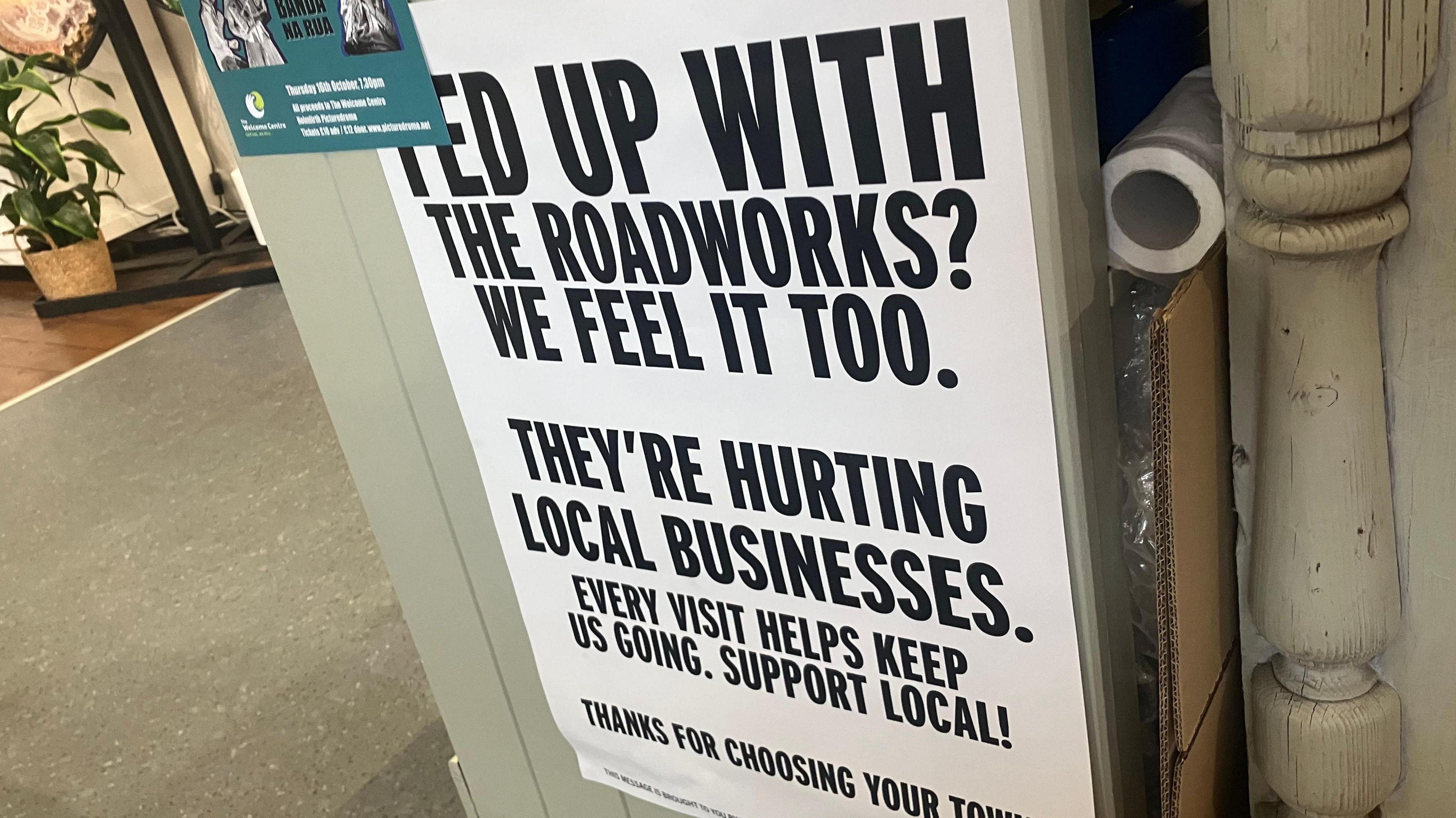 A black and white sign in a shop that reads: "Fed up with roadworks? We feel it too. They're hurting local businesses. Every visit helps keep us going. Support local!"