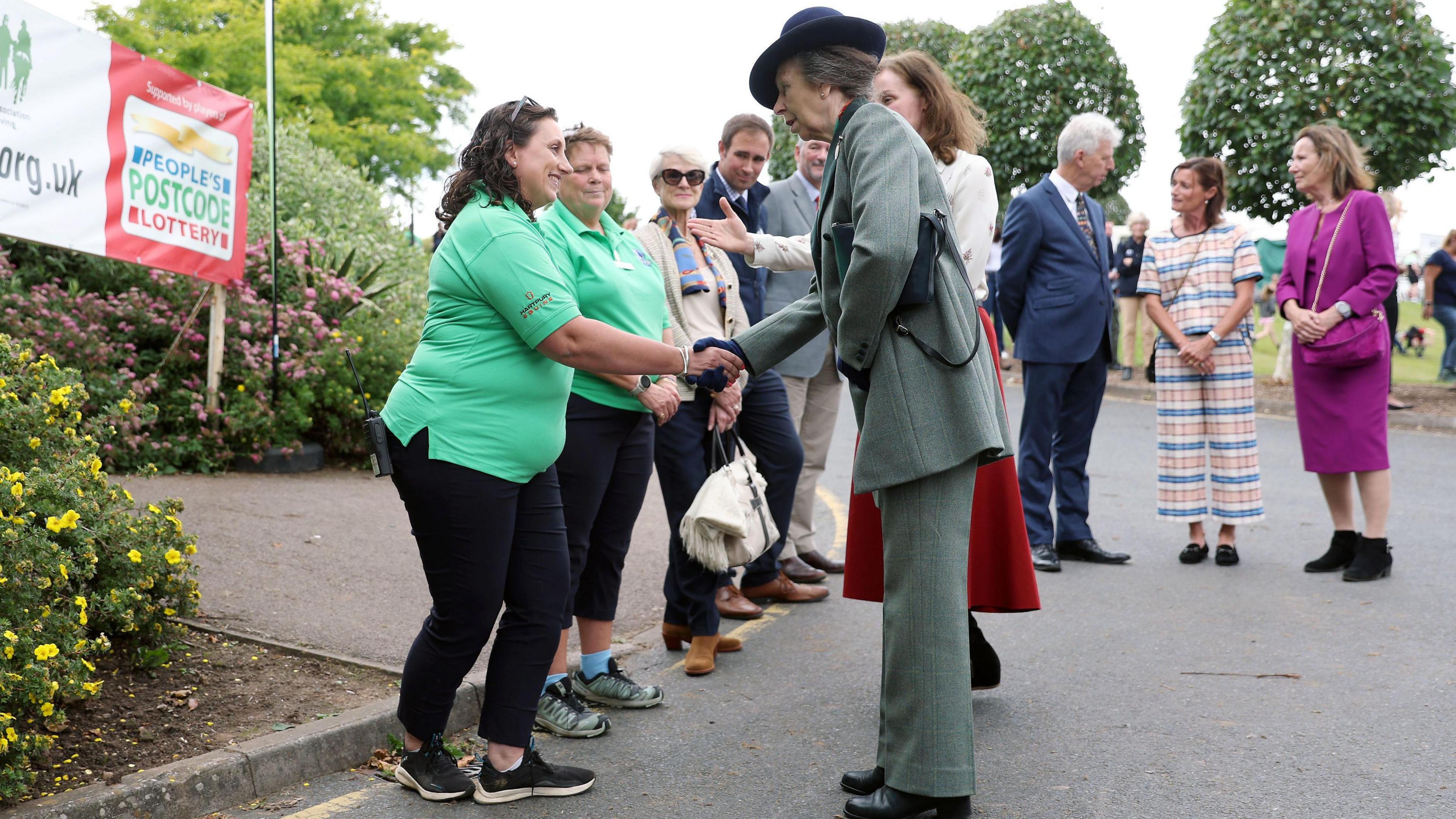 Princess Anne shaking hands with a woman. Princess Anne is wearing a green coat and trousers and a blue hat. The woman she is meeting has dark hair and a bright green top and black trousers on. 