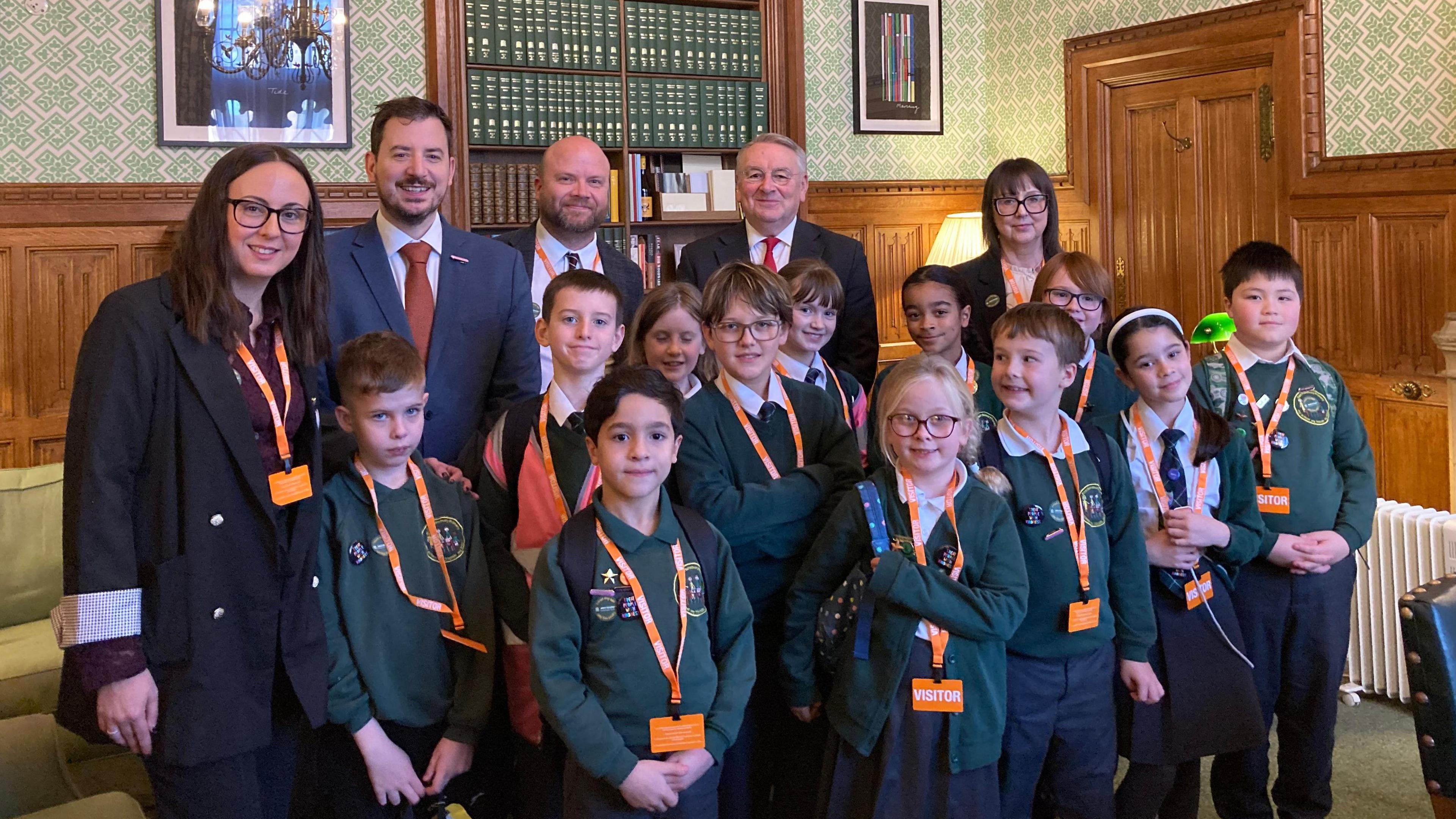 A group of schoolchildren and adults including MP Mike Reader and teacher Kelly Roberts pose in the House of Commons. The visitors are all wearing orange lanyards. The children are wearing green school jumpers.