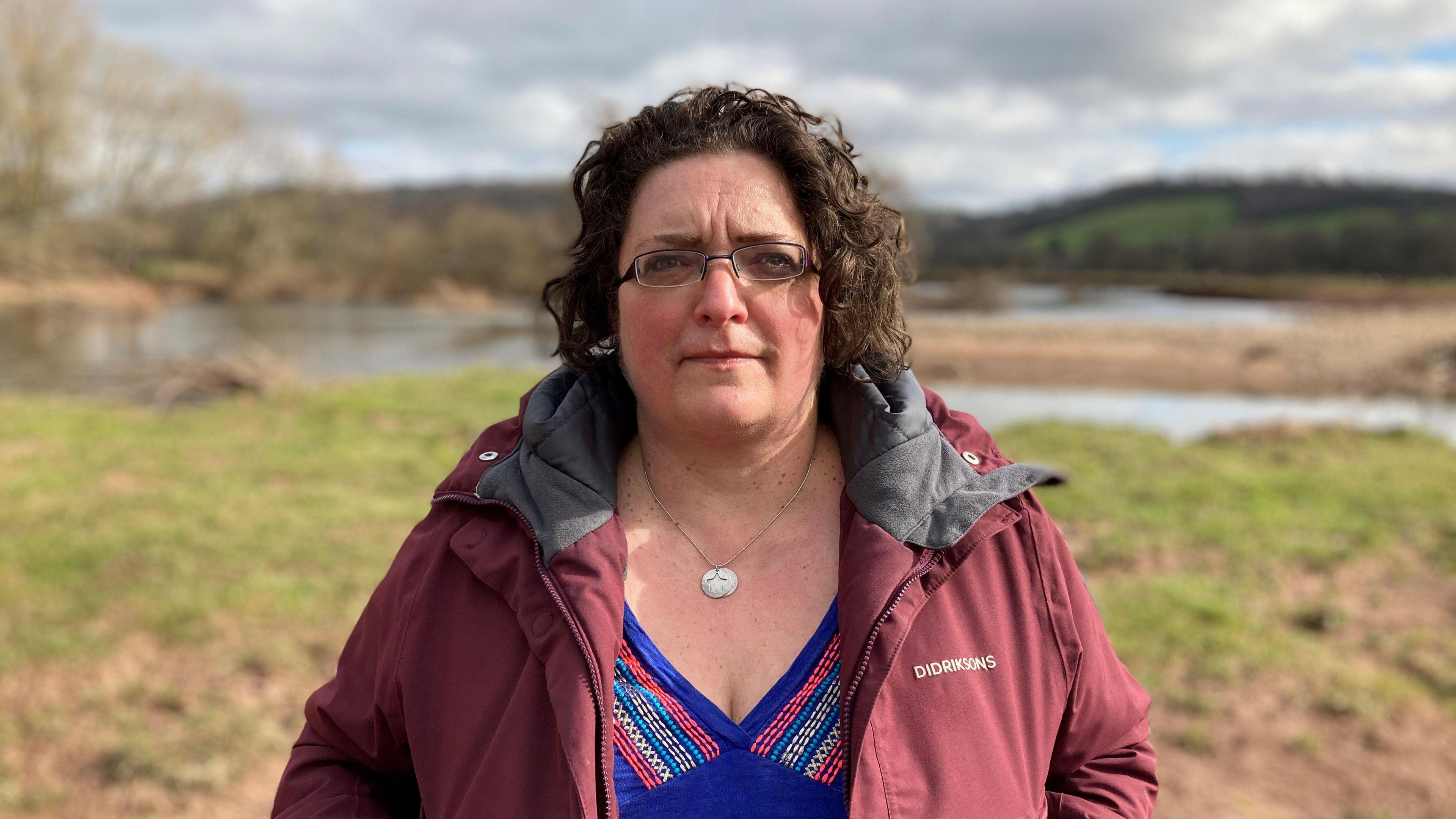 Gail Davies-Walsh, chief executive officer of Afonydd Cymru, which represents Wales' rivers trusts. She is standing on the banks of the river Usk near Newport, wearing a red coat and a blue top. She has brown curly hair and black glasses.