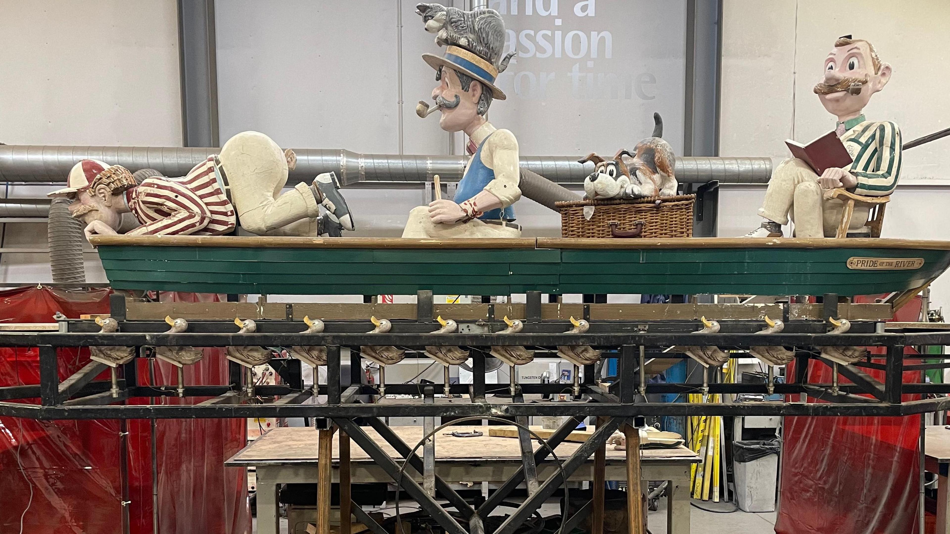 The mechanical workings of a boat sculpture are exposed. Within the boat are three cartoon-like mechanical figures who are male and in Edwardian dress, along with a sculpted dog which sits on top of a basket. One of the figures is kneeling and looking over the side of the boat, another is sitting upright on the boat, and the third is sitting on a small chair and appears to read from a book.