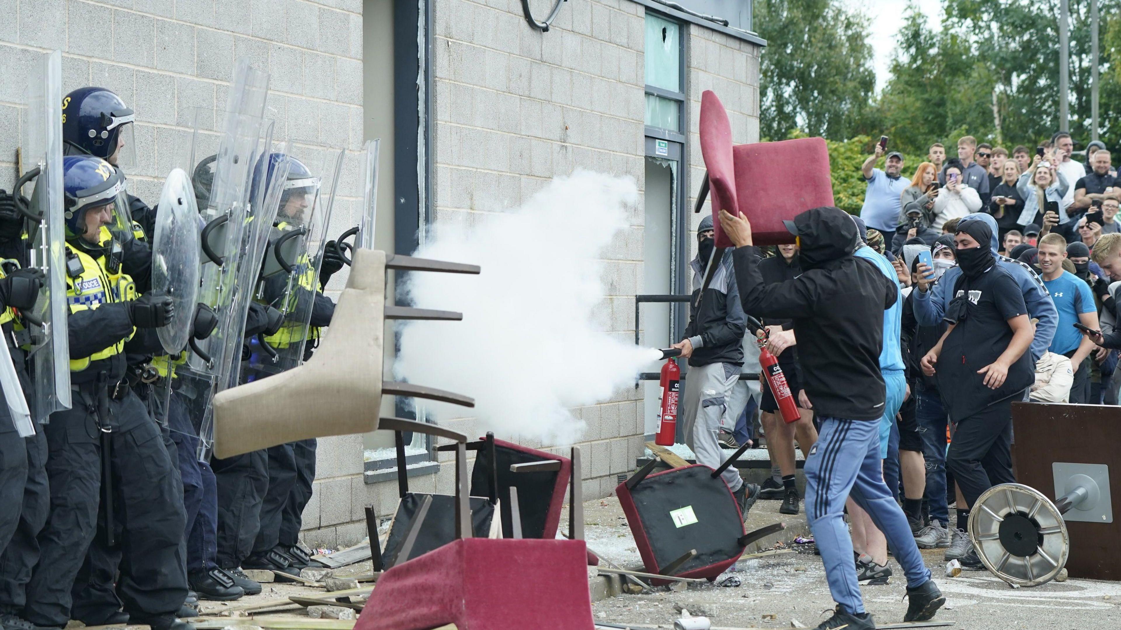 A man wearing blue tracksuit bottoms and a black hooded top throws a red chair at police officers.