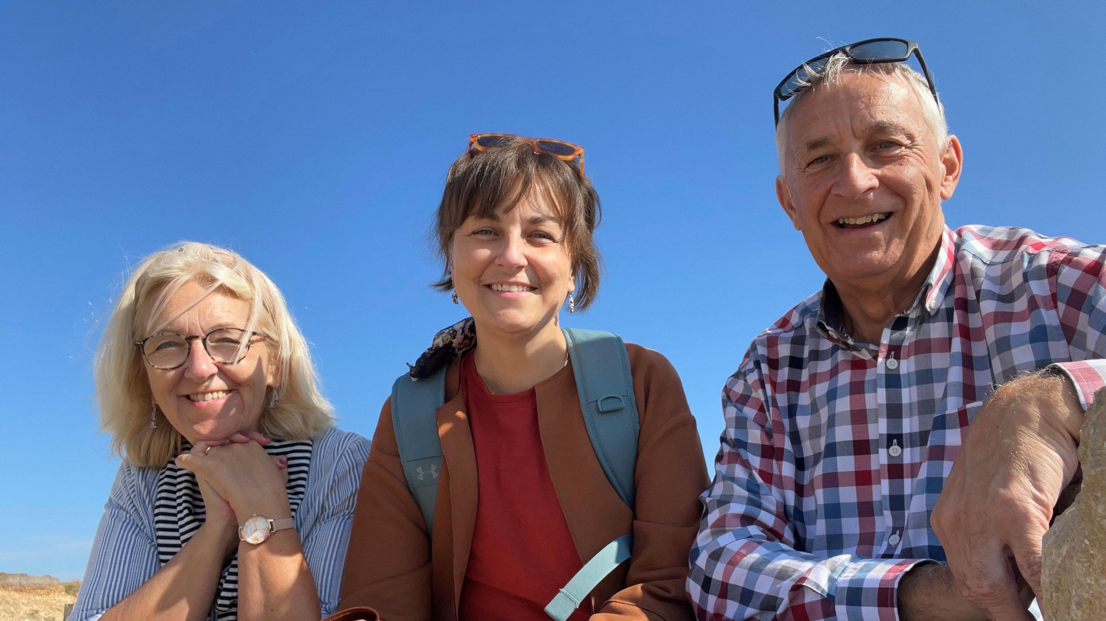 Julie, Sarah and John Wheldon sit in a row smiling for the camera against the backdrop of a bright blue sky. Julie, on the left, has shoulder length blonde hair, round glasses and a blue and white striped shirt. Sarah, who is in the middle, is wearing a brown coat and a blue rucksack. John, on the right, is wearing a large print gingham check shirt of blue, red and white. He has white hair and a pair of sunglasses on his head.