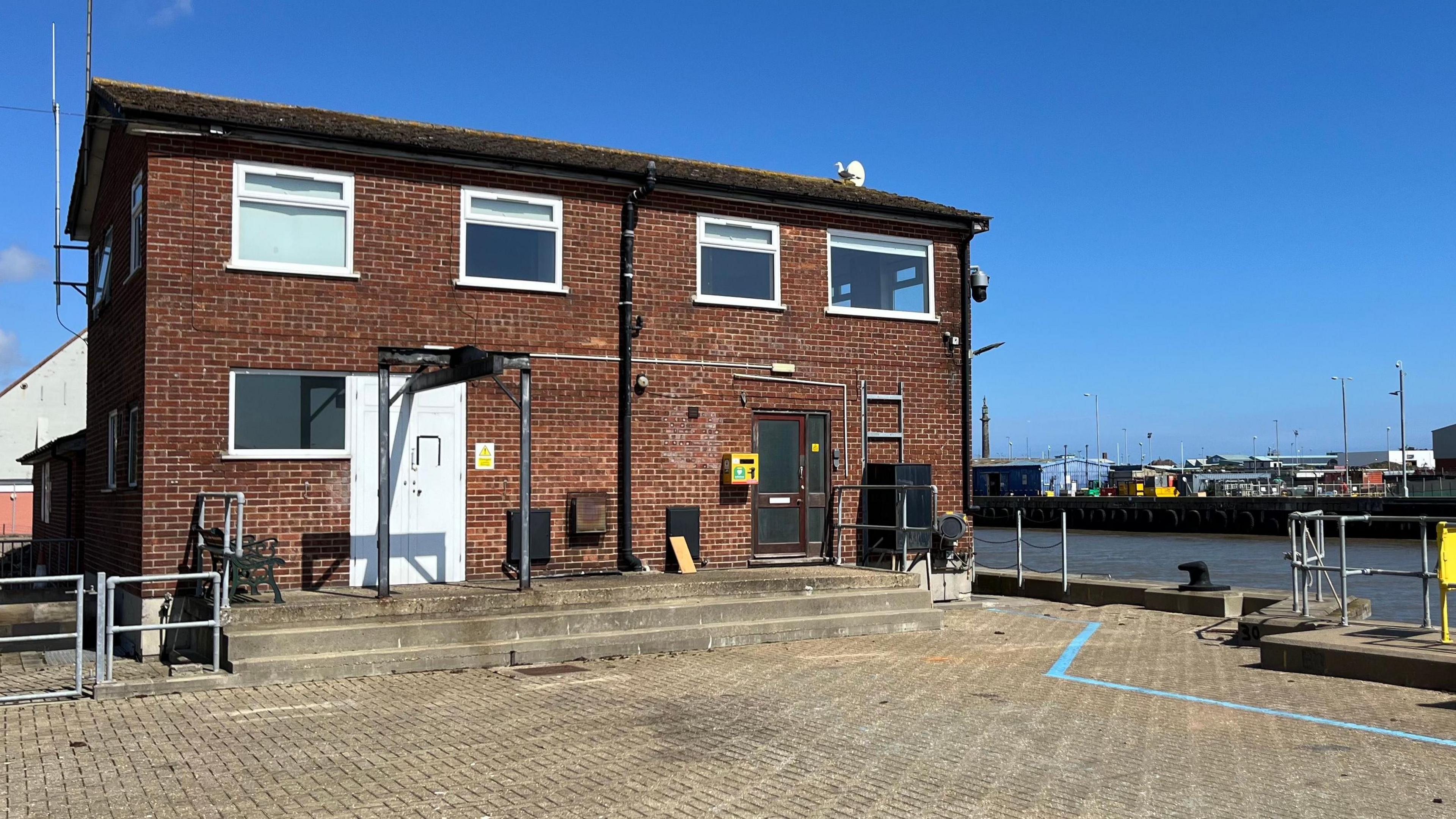 The Great Yarmouth River Port Pilots' Station in Gorleston is an red brick building of the 1970s, with a number of white-framed windows, and a shallow-pitched roof. In the foreground is brick-weave paving, with markings on the ground, and some hand rails and steps down to the river. Beyond the river, on the opposite back, are a series of port buildings and structures.