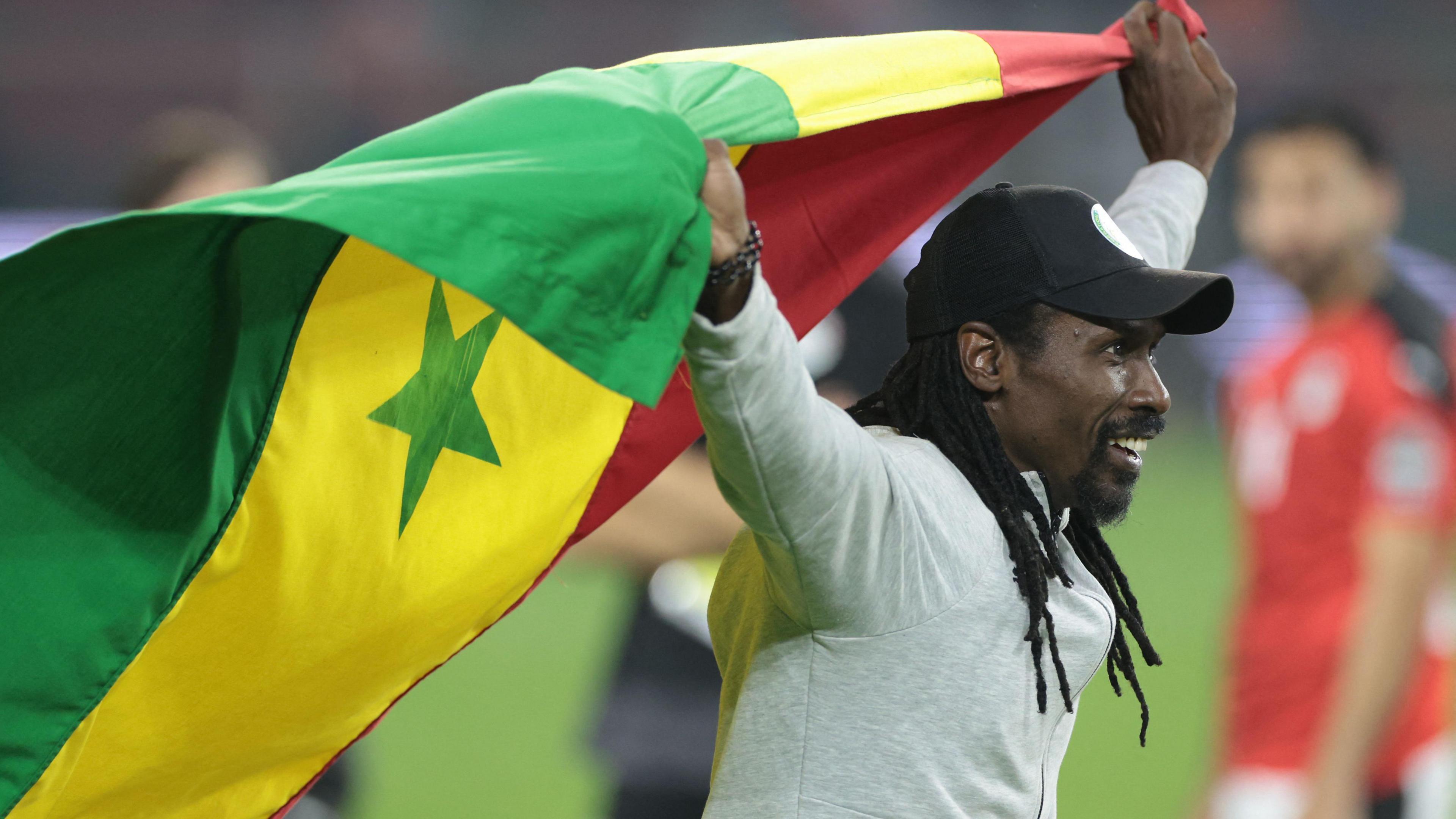 Aliou Cisse in grey top and black cap holds a Senegal flag aloft behind him as he celebrates winning the Africa Cup of Nations trophy