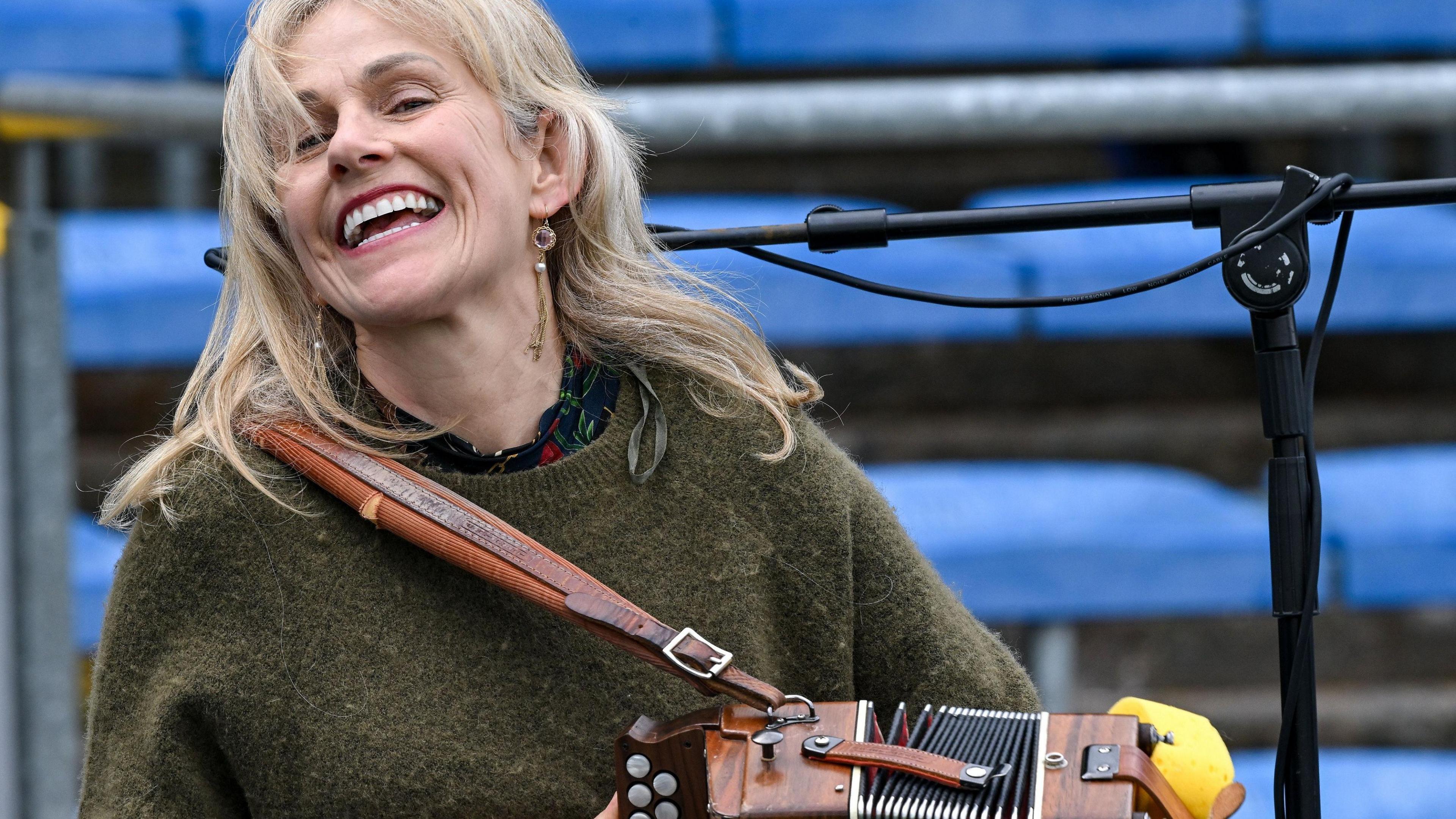 Sharon Shannon, a woman with long, blonde hair, smiles broadly as she plays a button accordion on stage. She is wearing a black, red and green top under an olive green jumper.