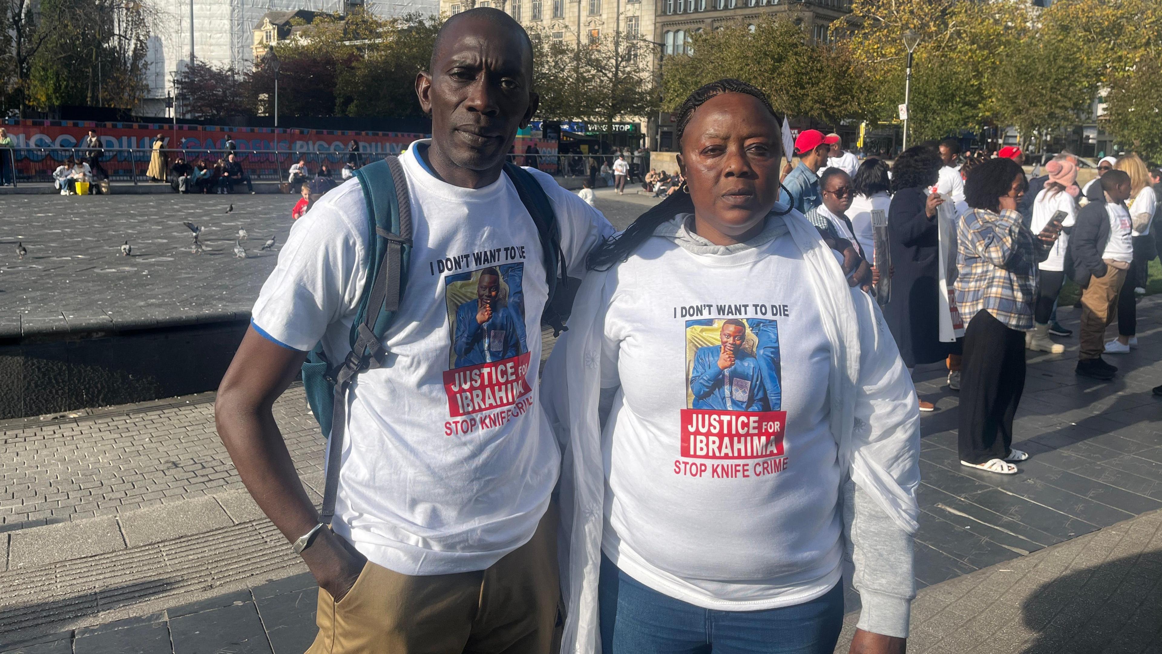 Mamadou Seck and Assa Ba stand together in Picadilly Gardens. They are both wearing T-shirts saying 'Justice for Ibrahima', 'I don't want to die' and 'Stop knife crime'. Other people are seen taking in the protest in the background.