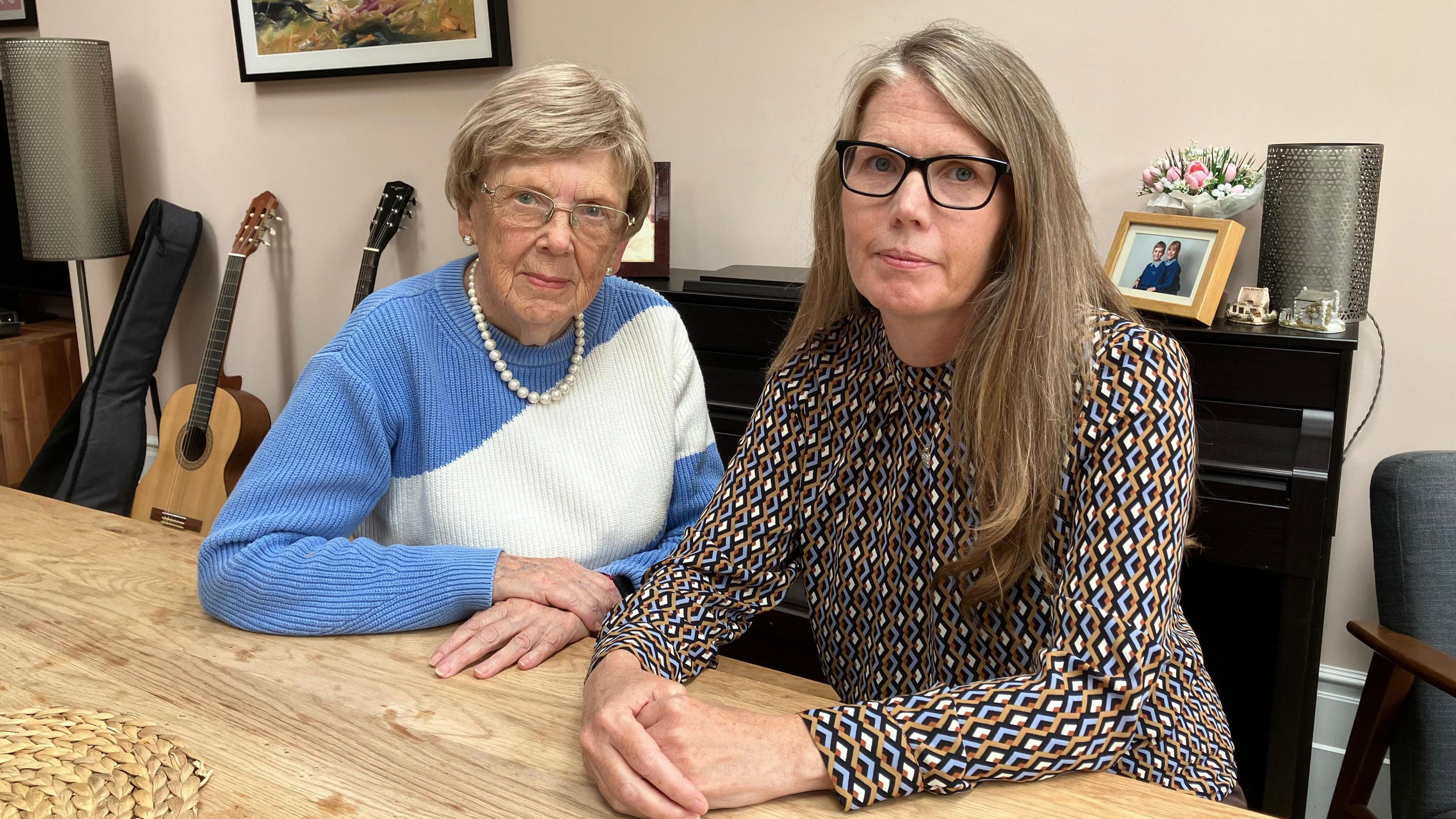 A woman sat on the left wearing a white and blue jumper and pearl necklace. A woman on the right is wearing a patterned top. They are sat at a wooden table.