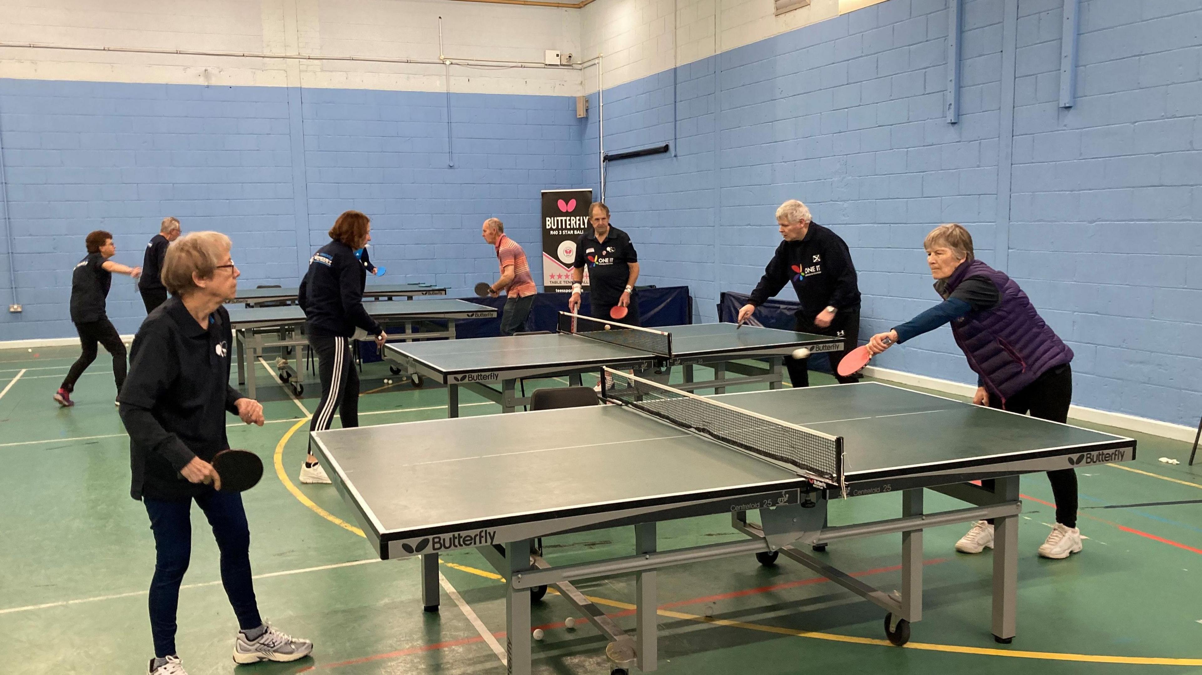 Four green table tennis tables next to each other with a pair of adults on each table playing each other. They are all largely wearing black clothing. They are in a hall with blue walls and green flooring.