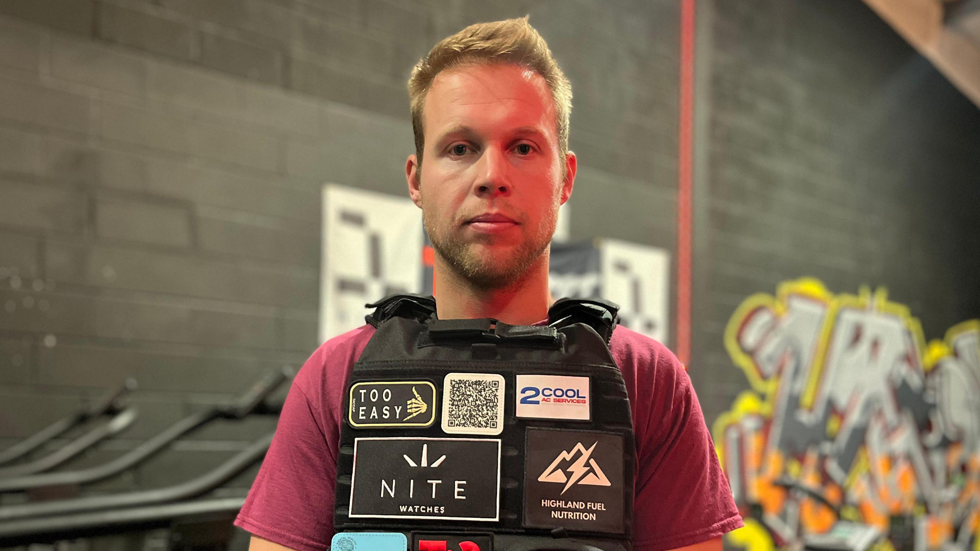 A man wearing a burgundy t-shirt and a black vest is pictured stood in front of a black painted wall. 