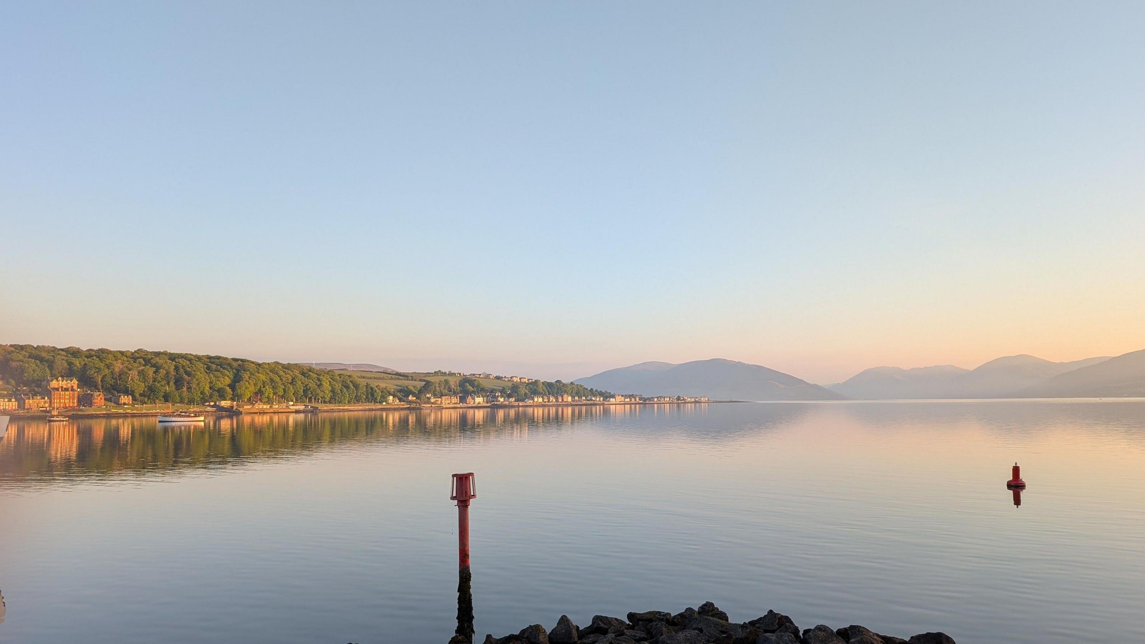 A calm body of water with two buoys, a rocky shore in the foreground, distant hills, and a clear sky at sunrise or sunset.