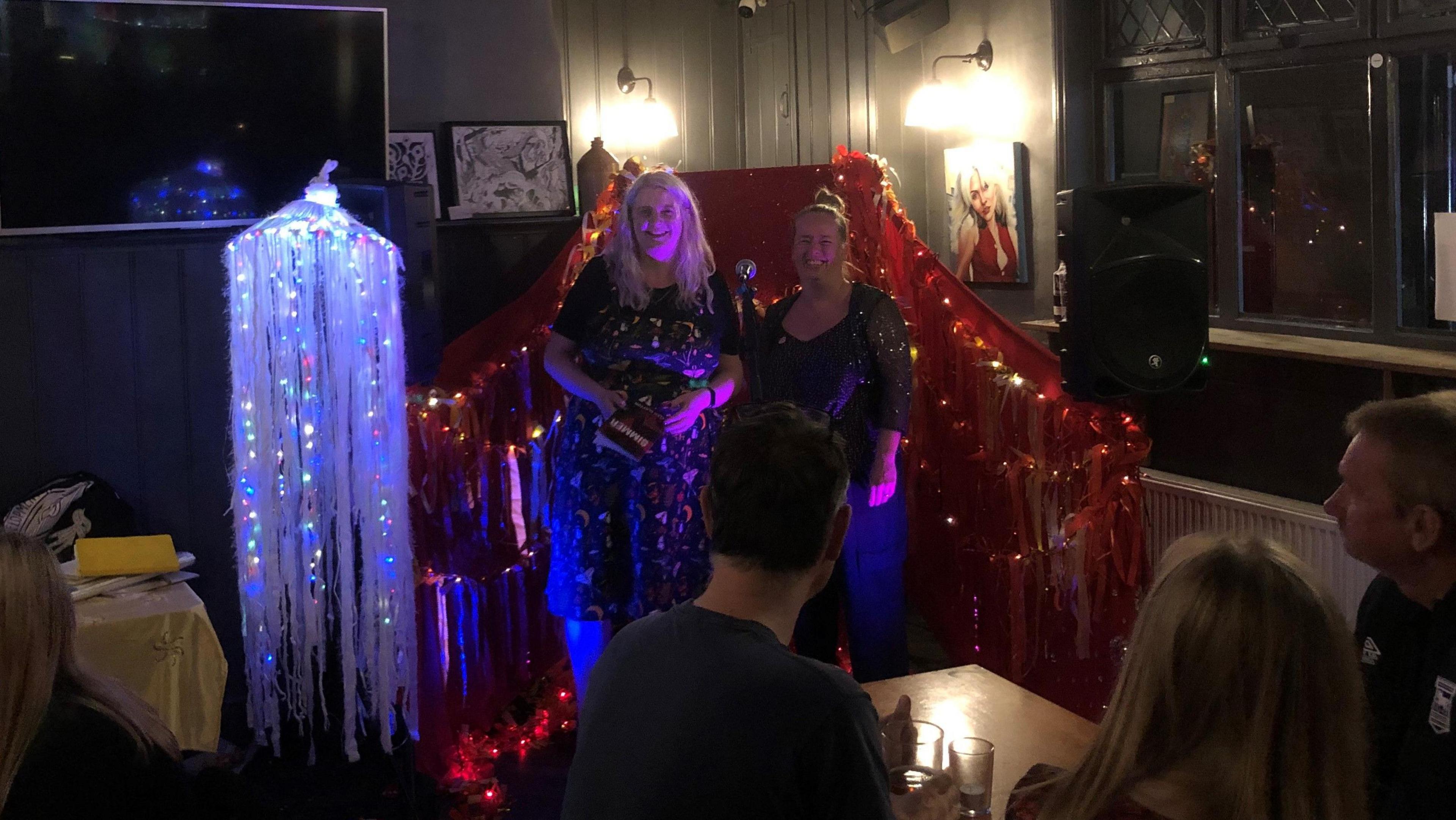 Ellen Clayton with poet and promotor Amy Wragg standing by a microphone on a stage in a pub. The stage is decorated with lights and flowing material. In the foreground there are people sitting at tables.