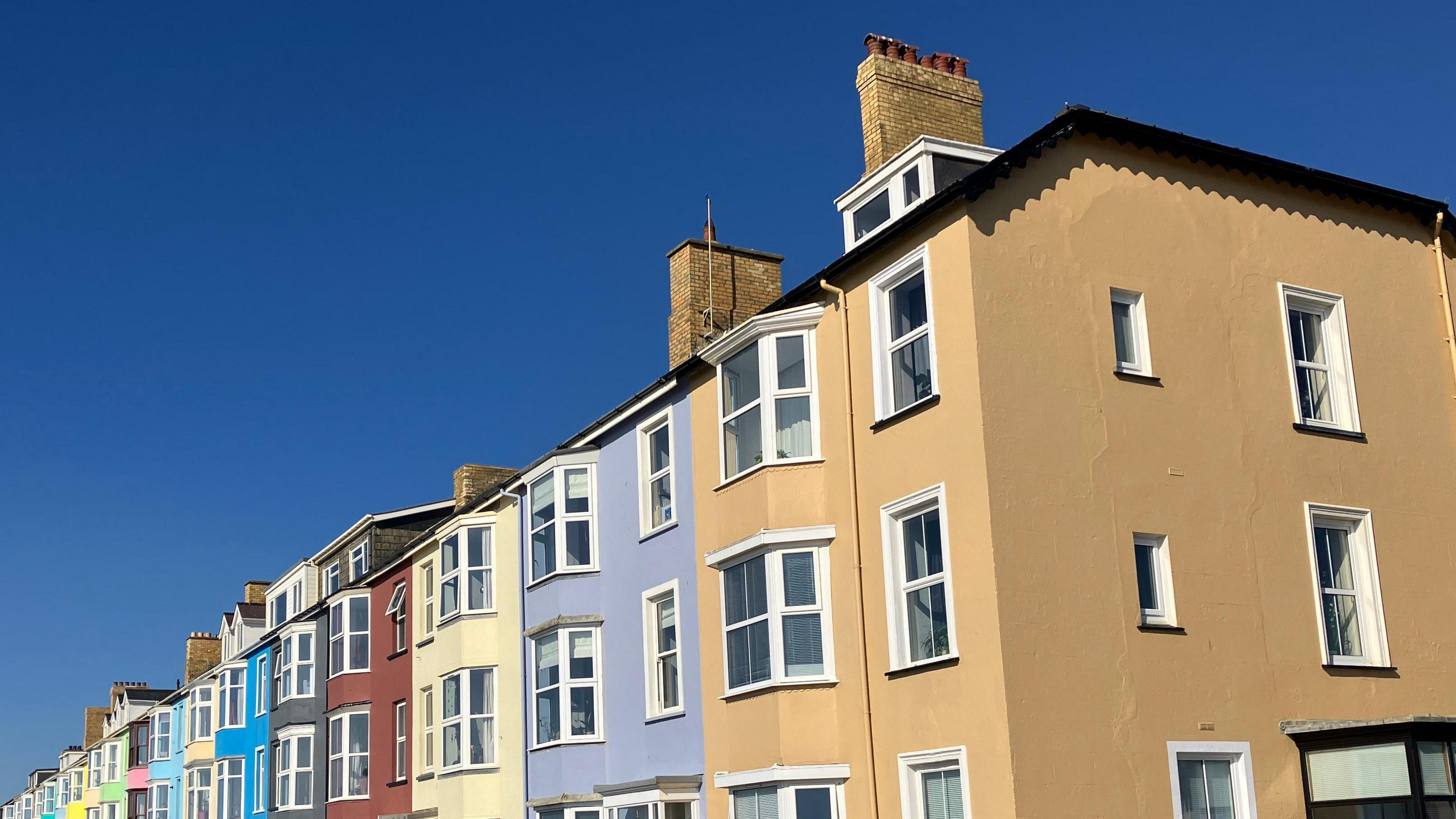 A street in Aberystwyth. A row of colourful townhouses with white window sills sit side by side, in colours like yellow, purple, red, grey, and blue. The sky behind them is bright blue. 