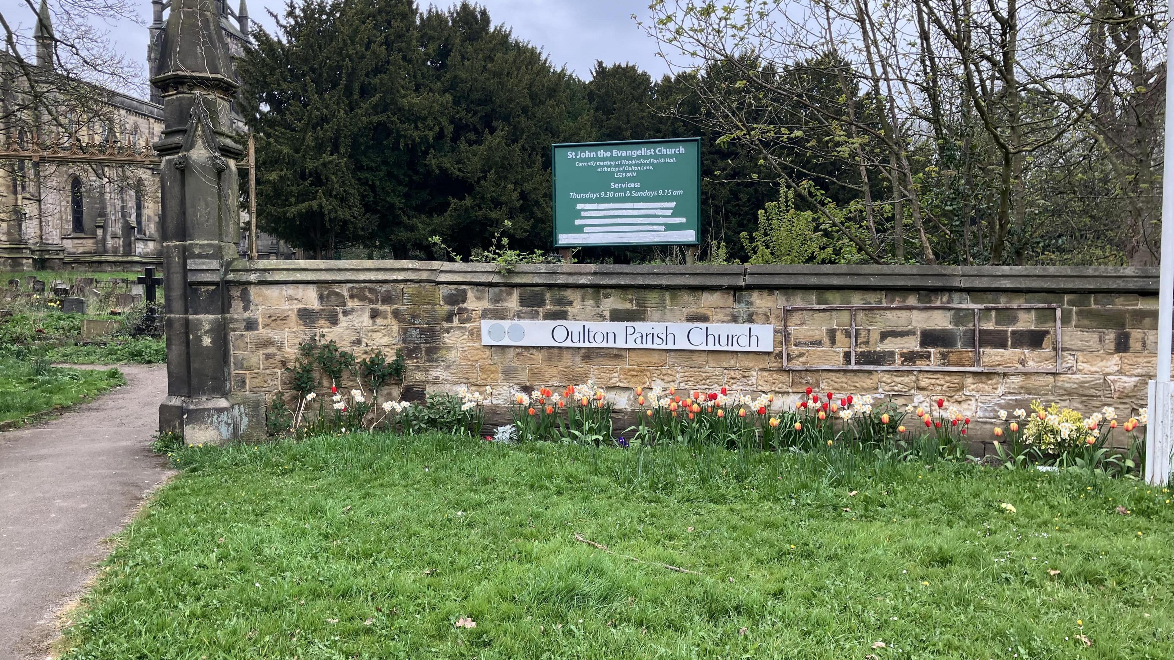 The boundary wall of a church, bearing the sign 'Oulton Parish Church'. Some flowers are at the foot of the wall at the edge of a patch of grass.