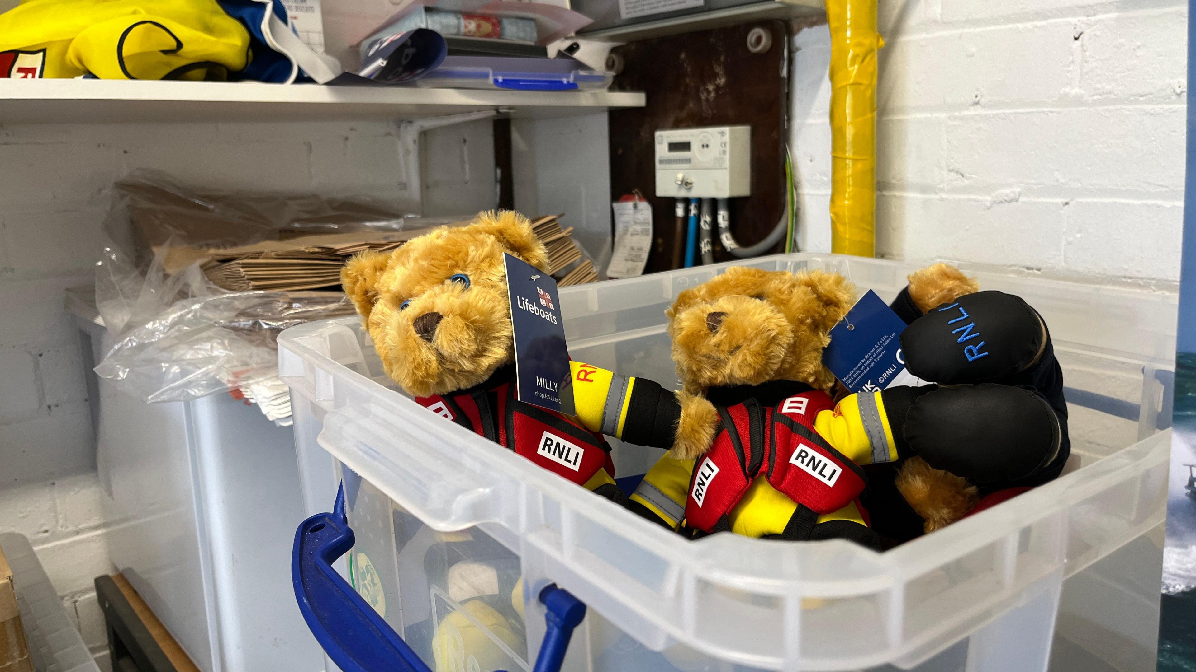 A white plastic box inside the shop that has been filled with little brown teddy bears. They are wearing red RNLI life jackets and a white high vis underneath. 