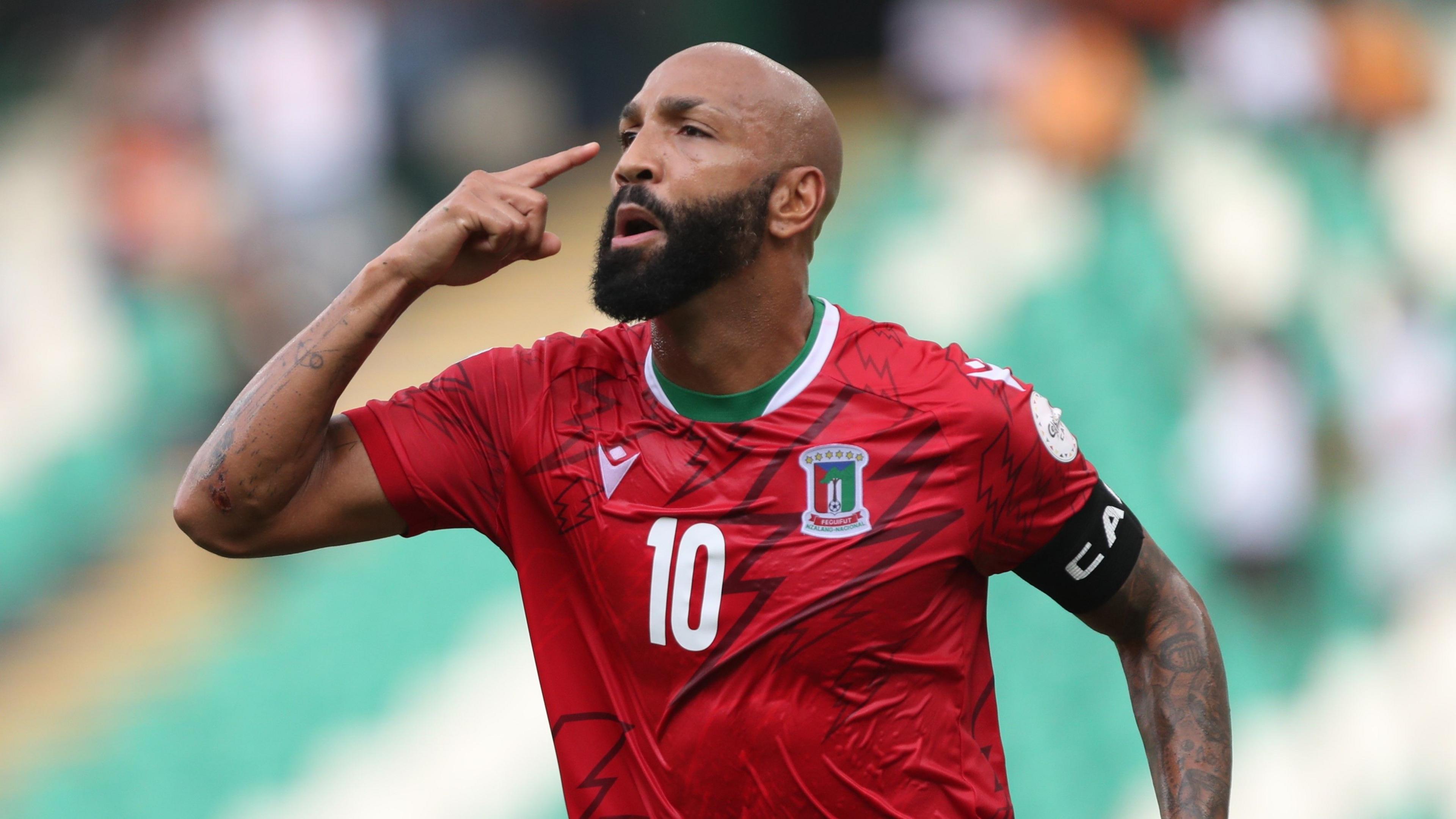 Emilio Nsue in red Equatorial Guinea shirt and with captain's armband points with his right finger at his temple.