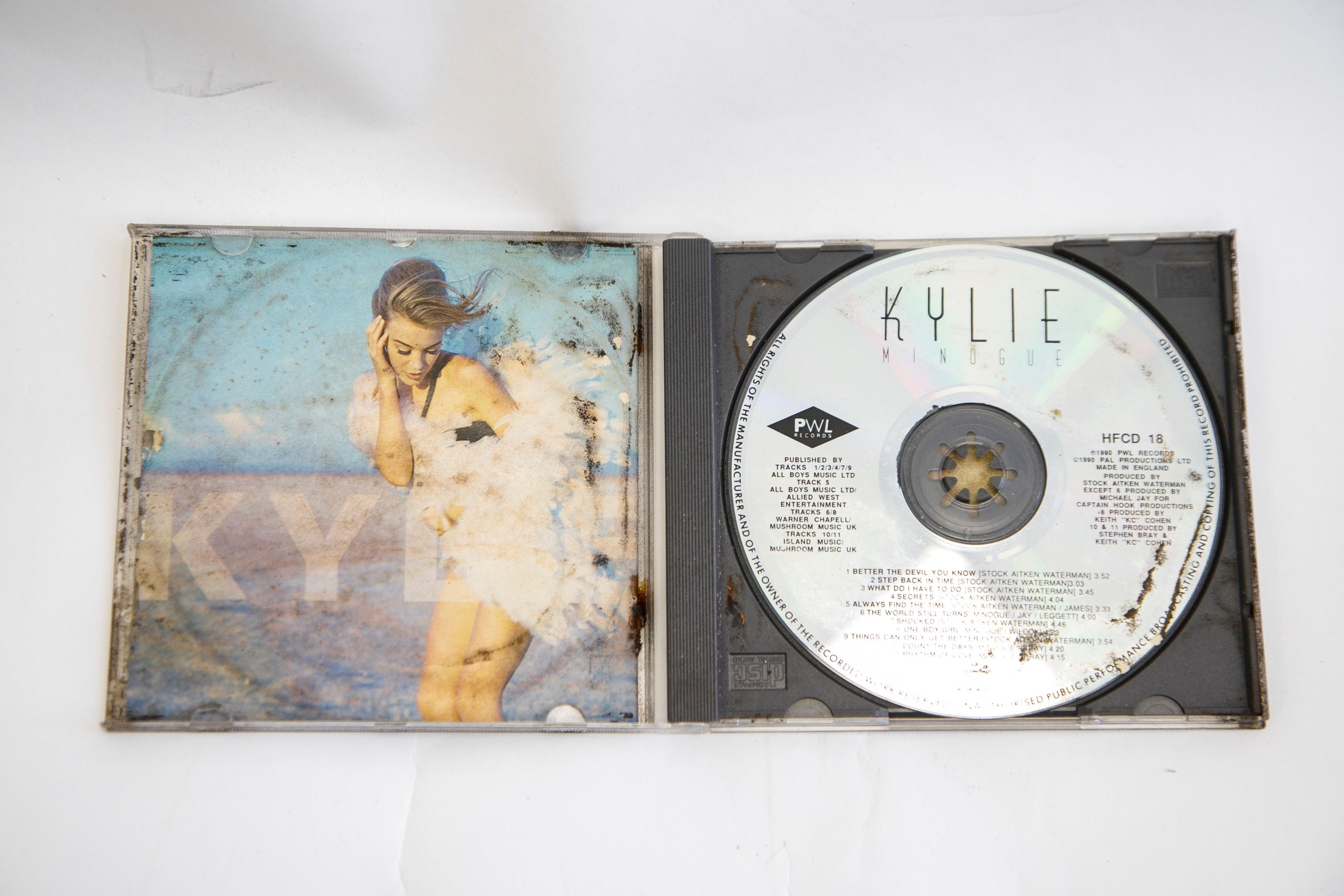 The CD of Kylie Minogue's album Rhythm of Love is open on a table. There is rust and dirt on different areas of the CD, which is metallic silver in colour. 