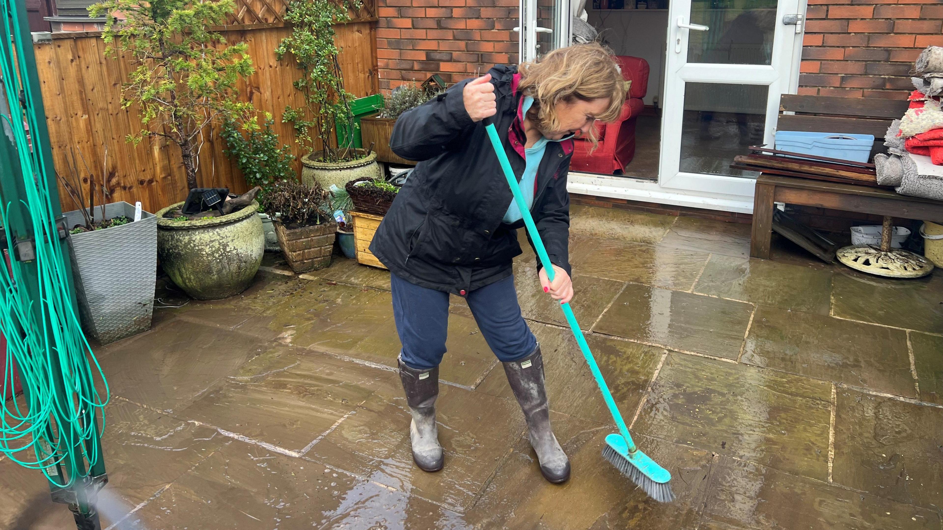 Bernadette Melea, 70, who has blonde hair and wears a black coat, brown wellington boots and jeans, sweeps water from her paved back yard