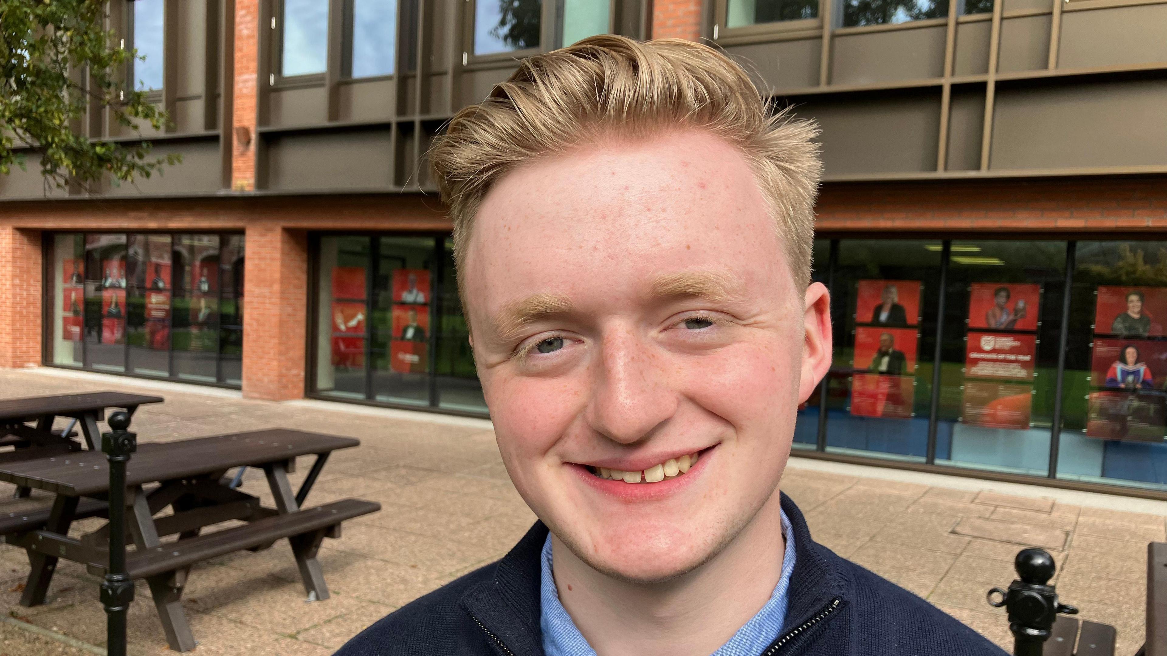 Oliver is standing in front of a modern glass red brick building and picnic benches. 

He has blue eyes, fair eyebrows and short hair pushed back. 

Oliver is wearing a blue shirt and navy fleece over it. 

He is smiling at the camera. 