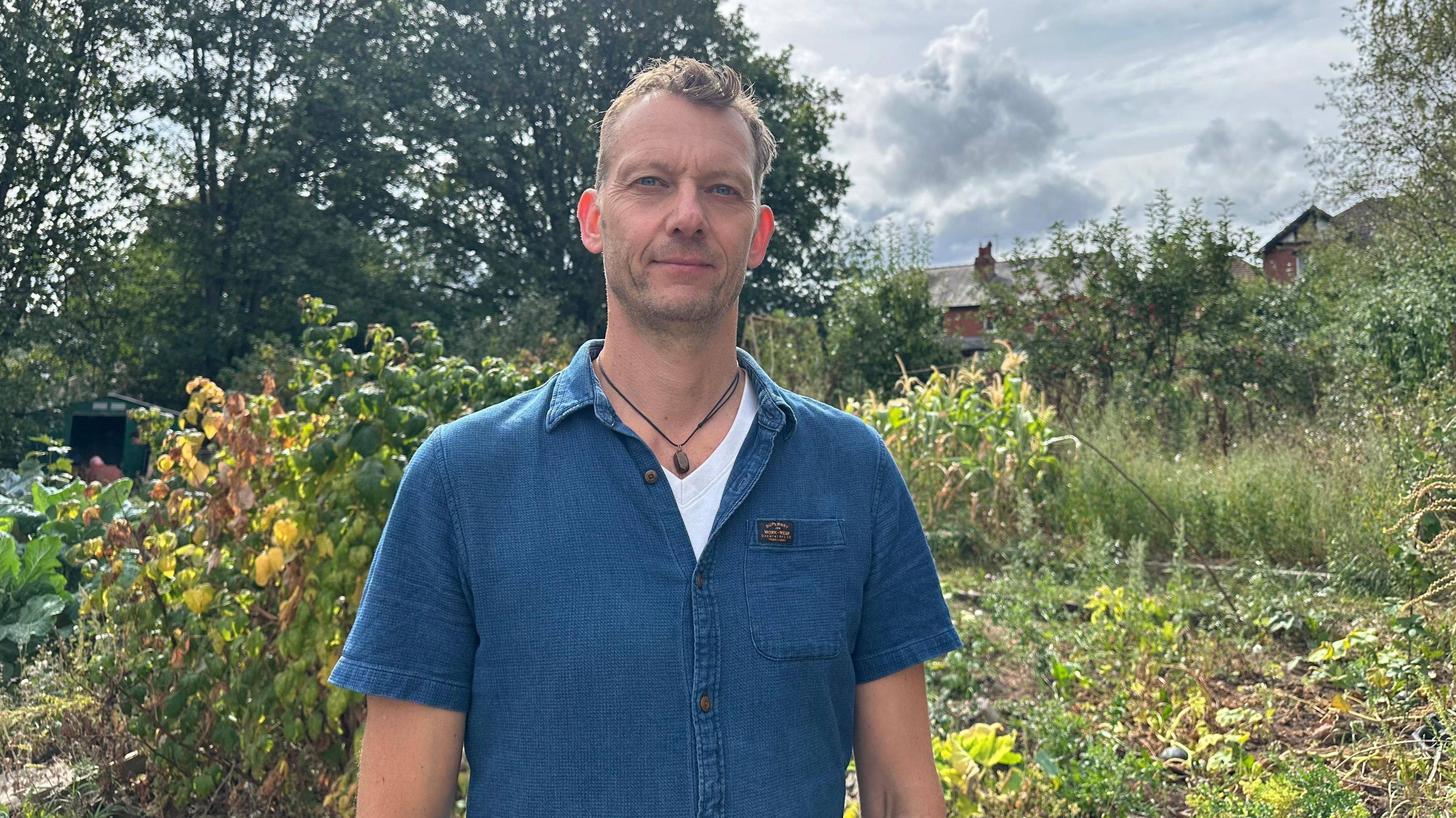 Professor Paul Chatterton is standing in the allotment site at LILAC community housing scheme, in Leeds. He is wearing a blue shirt and is looking straight at the camera.