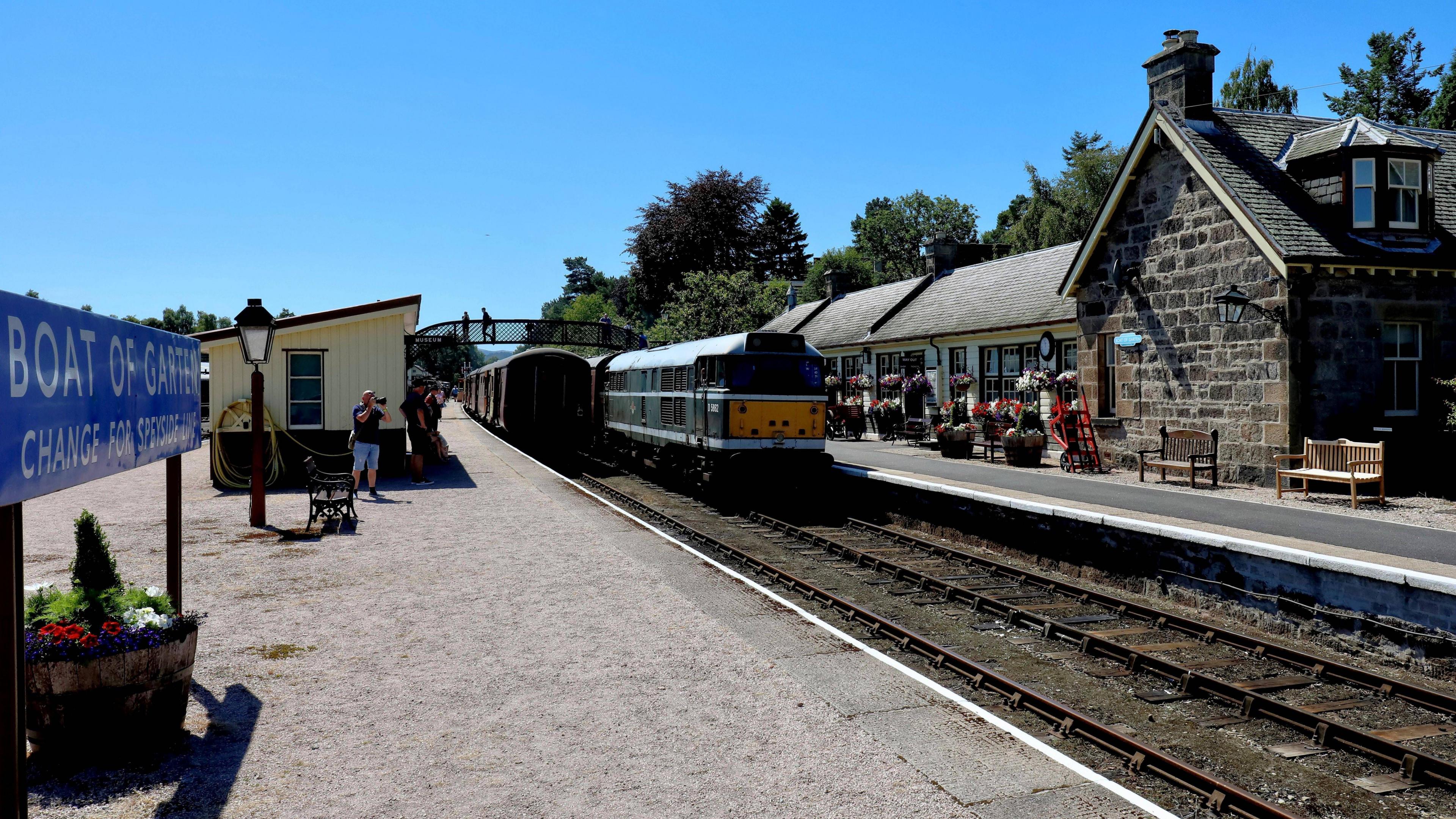 A heritage diesel train rolls into a train on a very sunny day, watched by a few men with cameras