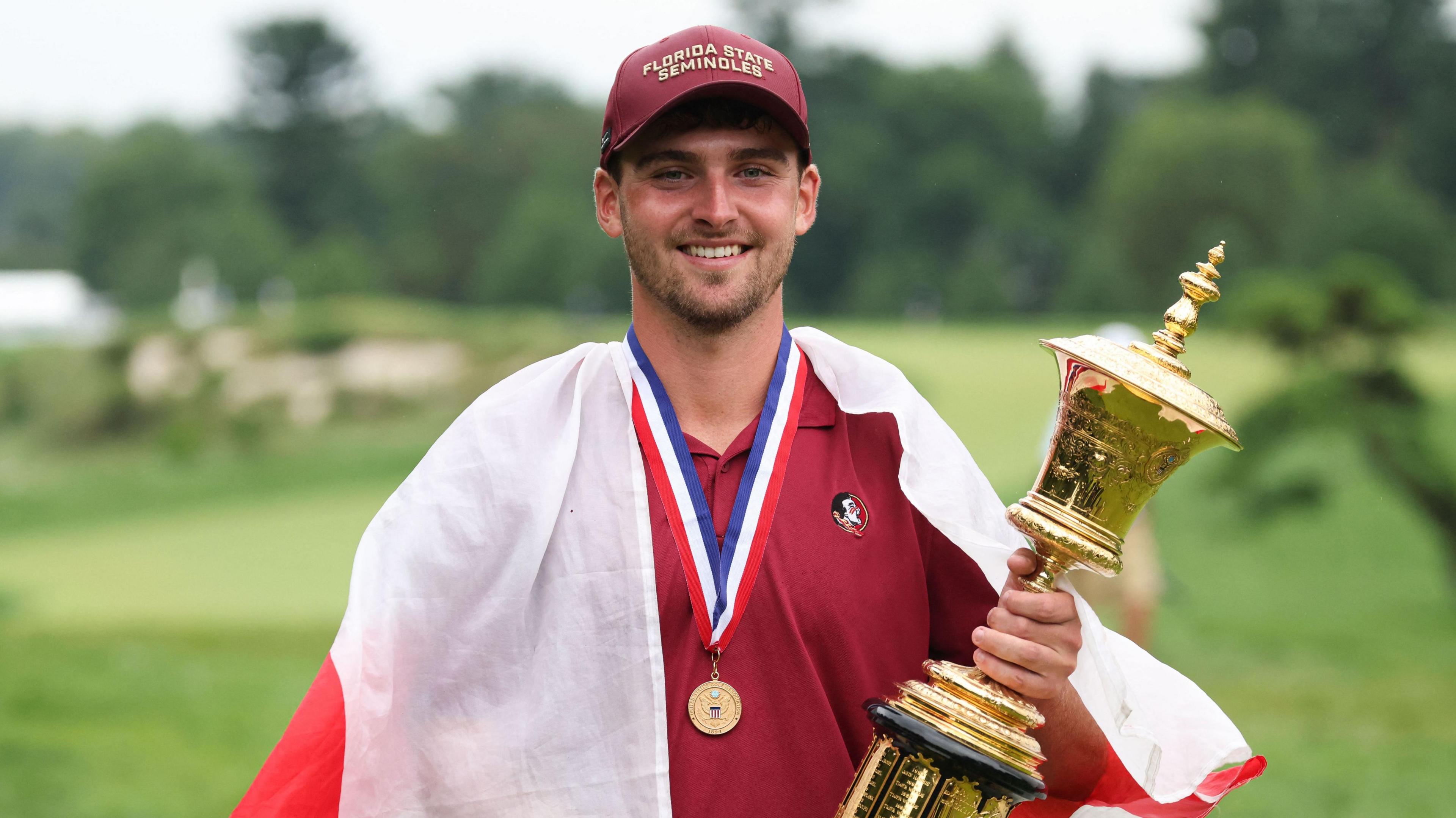 Jack Whaley holding the US Amateur trophy in his left hand with a medal around his neck and an England flag draped around his shoulders