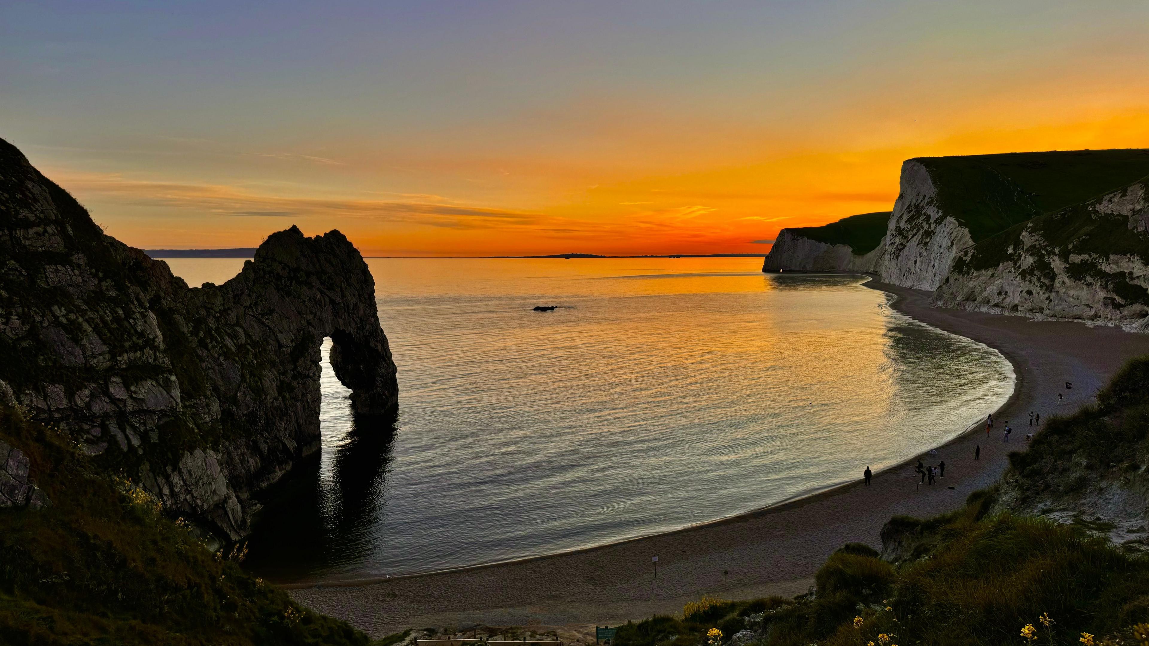 A sunset image of Lulworth Cove with its curved beach and high cliffs including Durdle Door. The sky is a brilliant hue of orange fading through to blues over the calm sea