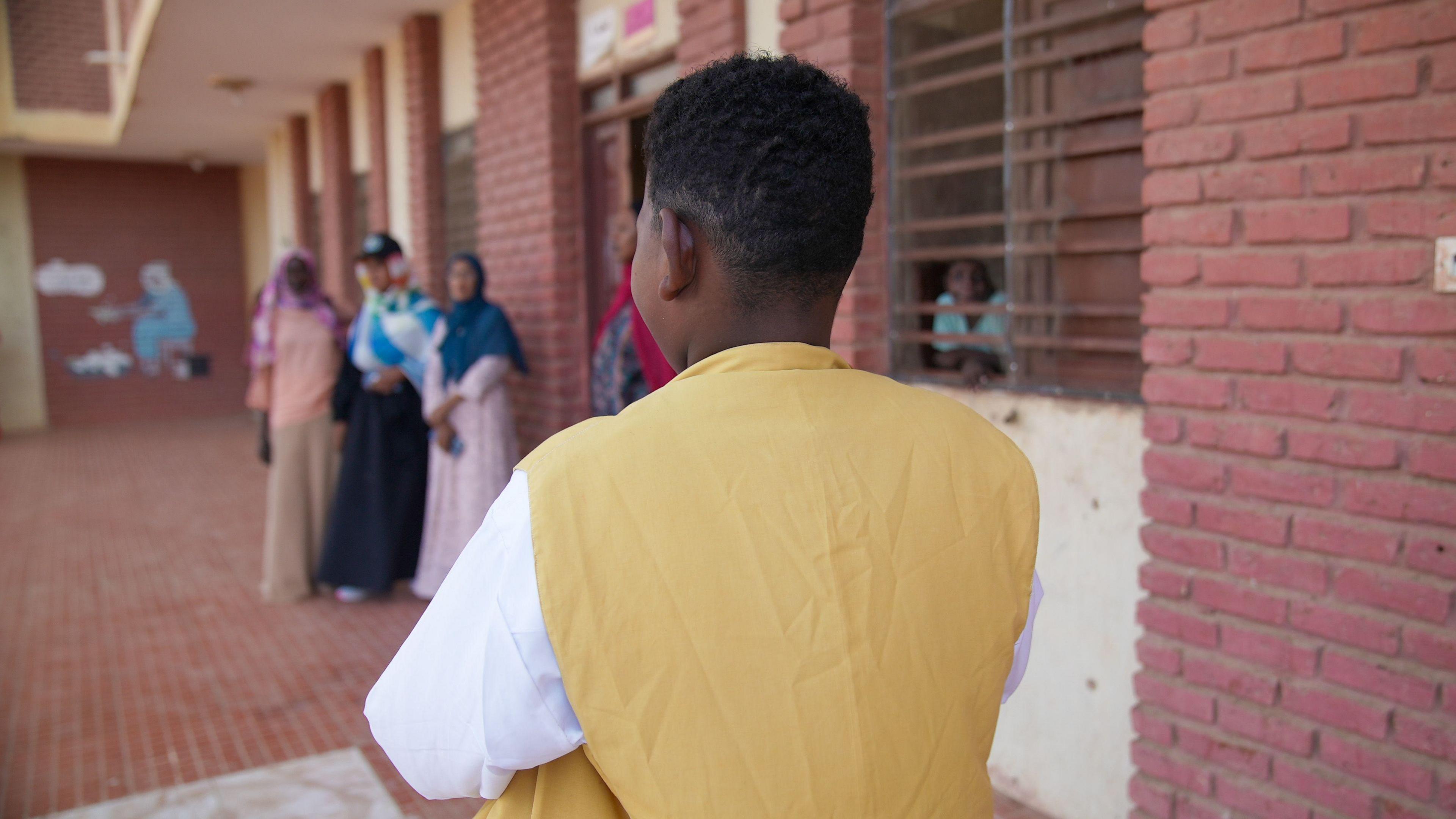 The back of Mahmoud, a teenager in a yellow and white top, standing next to a building with three women stood in the distance, just out of focus