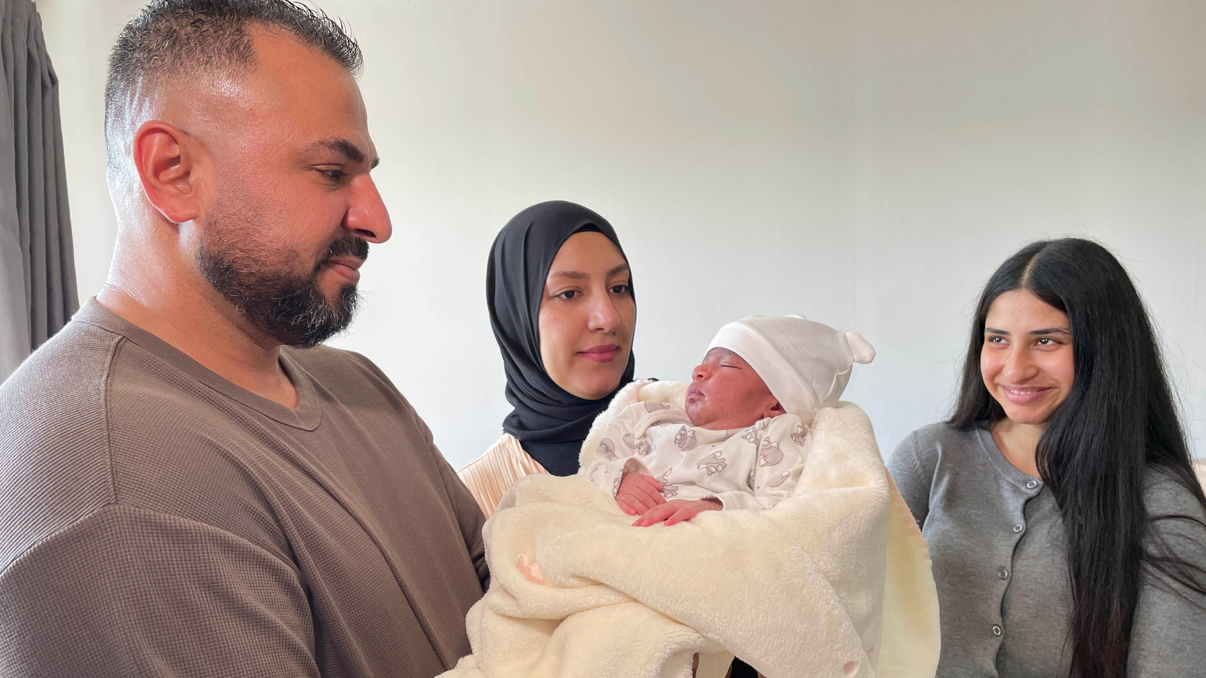 Ahmed with his wife, Nour and teenage daughter, Rahaf. They are all happy and focused on a sleeping newborn baby, being held by Ahmed and wrapped in a cream coloured blanket.   