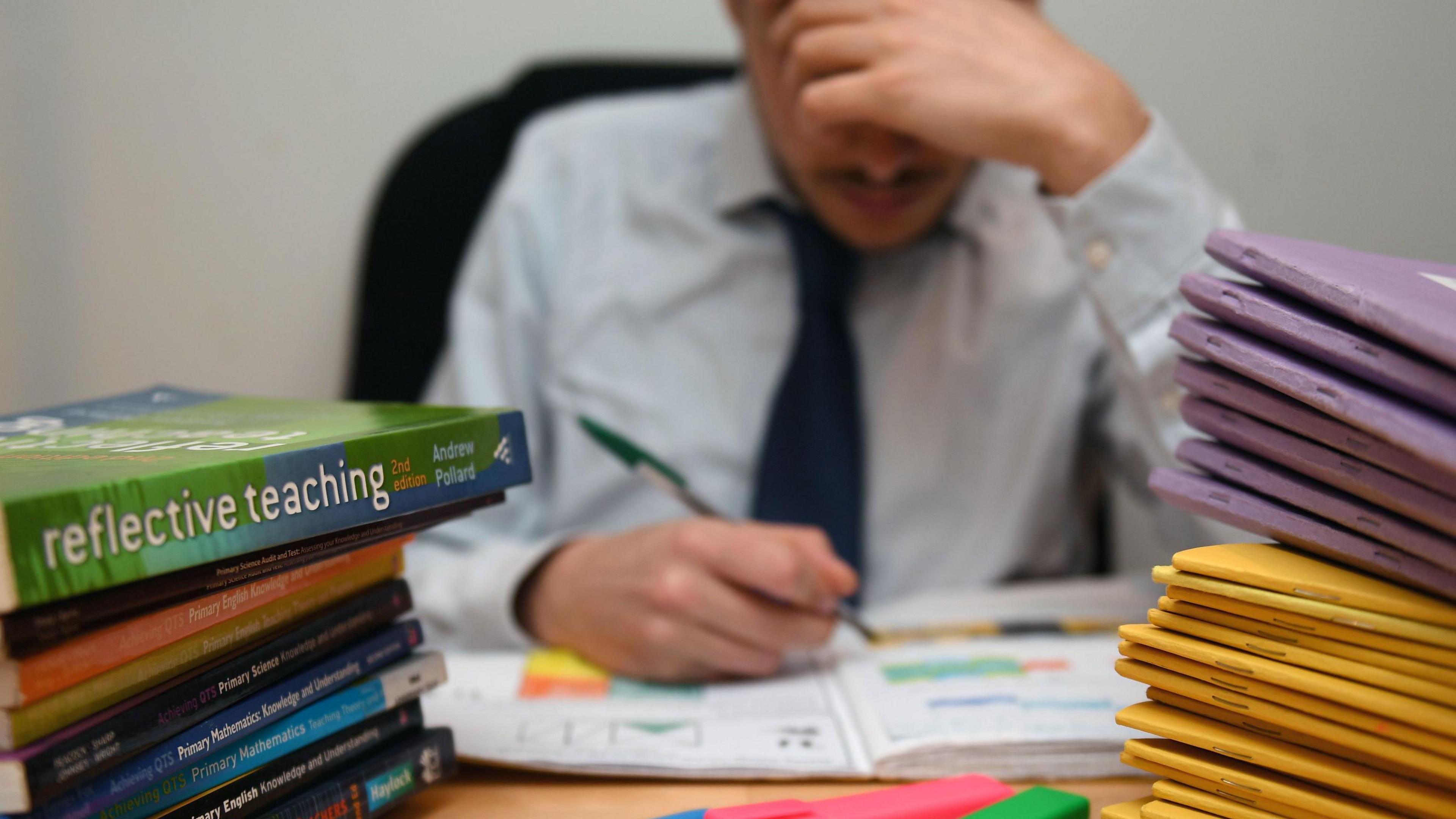 Teenage boy in shirt and school tie writing in a workbook at a desk with pens and other work books piled on it
