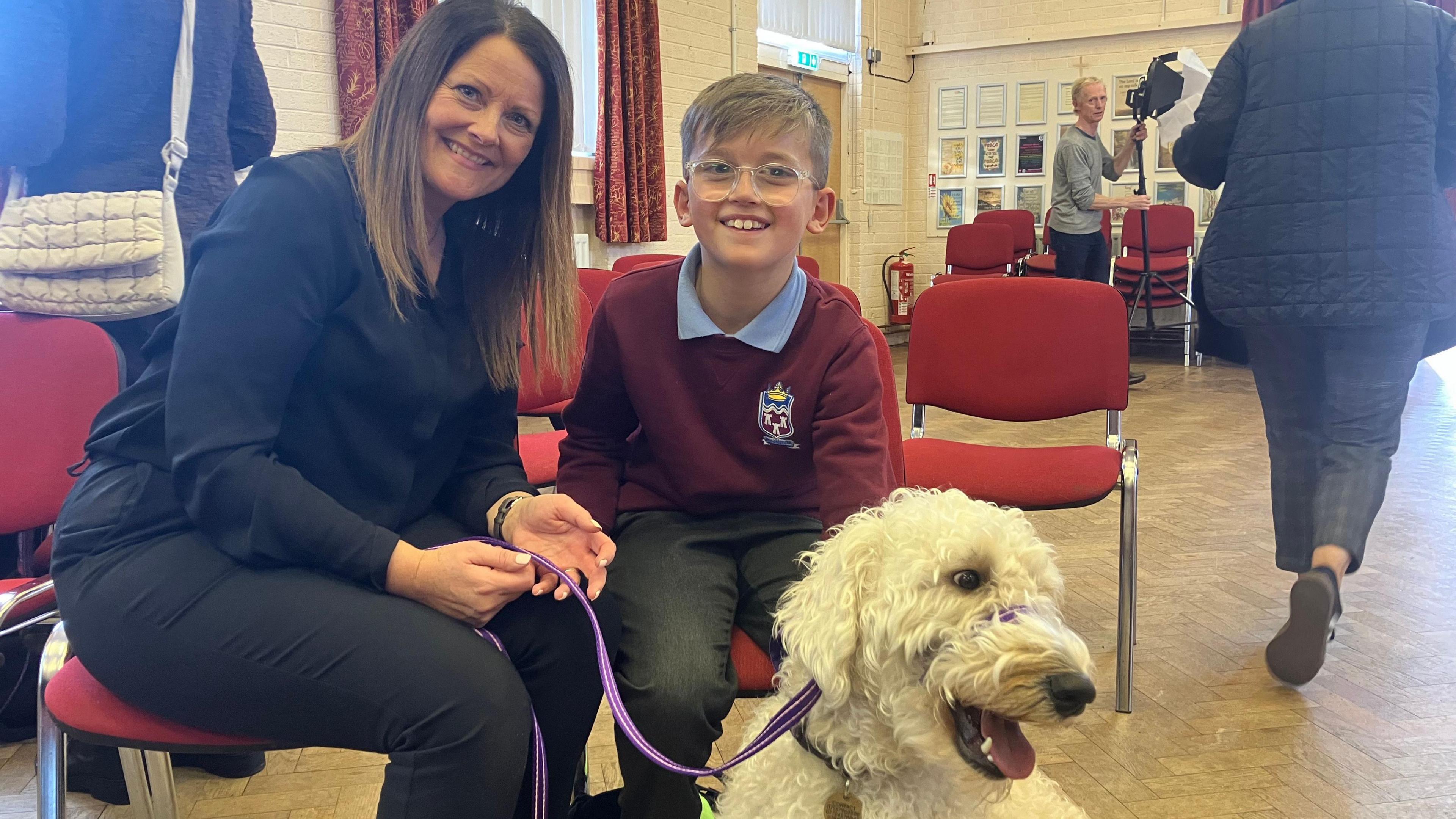 Fiona Smart is wearing a black shirt and black trousers. Caleb, who is a boy sitting next to hear,  is wearing his purple school jumper, with a blue t-shirt, and grey trousers. Peanut, who's a white labradoodle, is looking to the side, with their tongue sticking out.