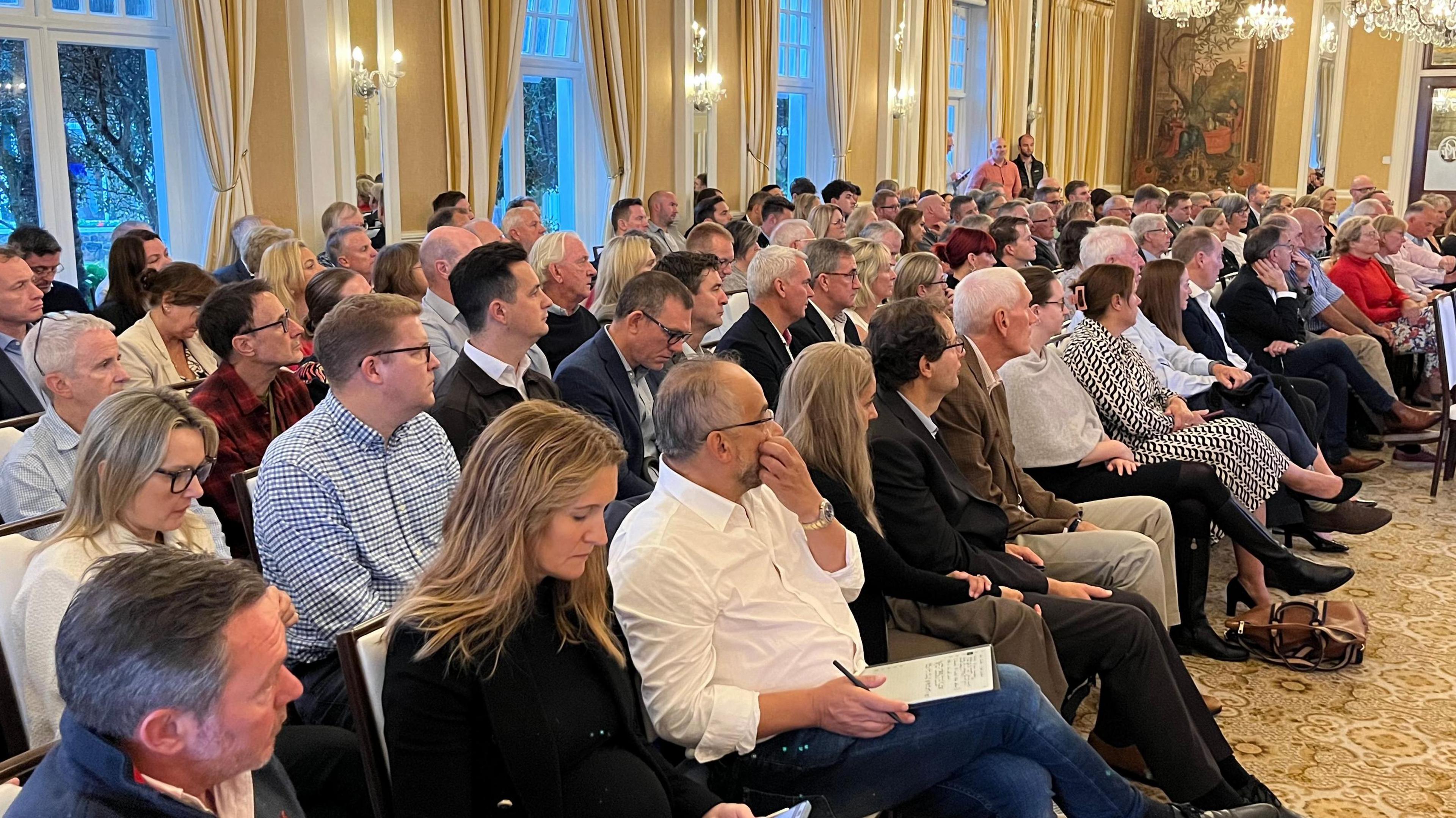 A group of about 100 people sitting in a room with chandeliers. 