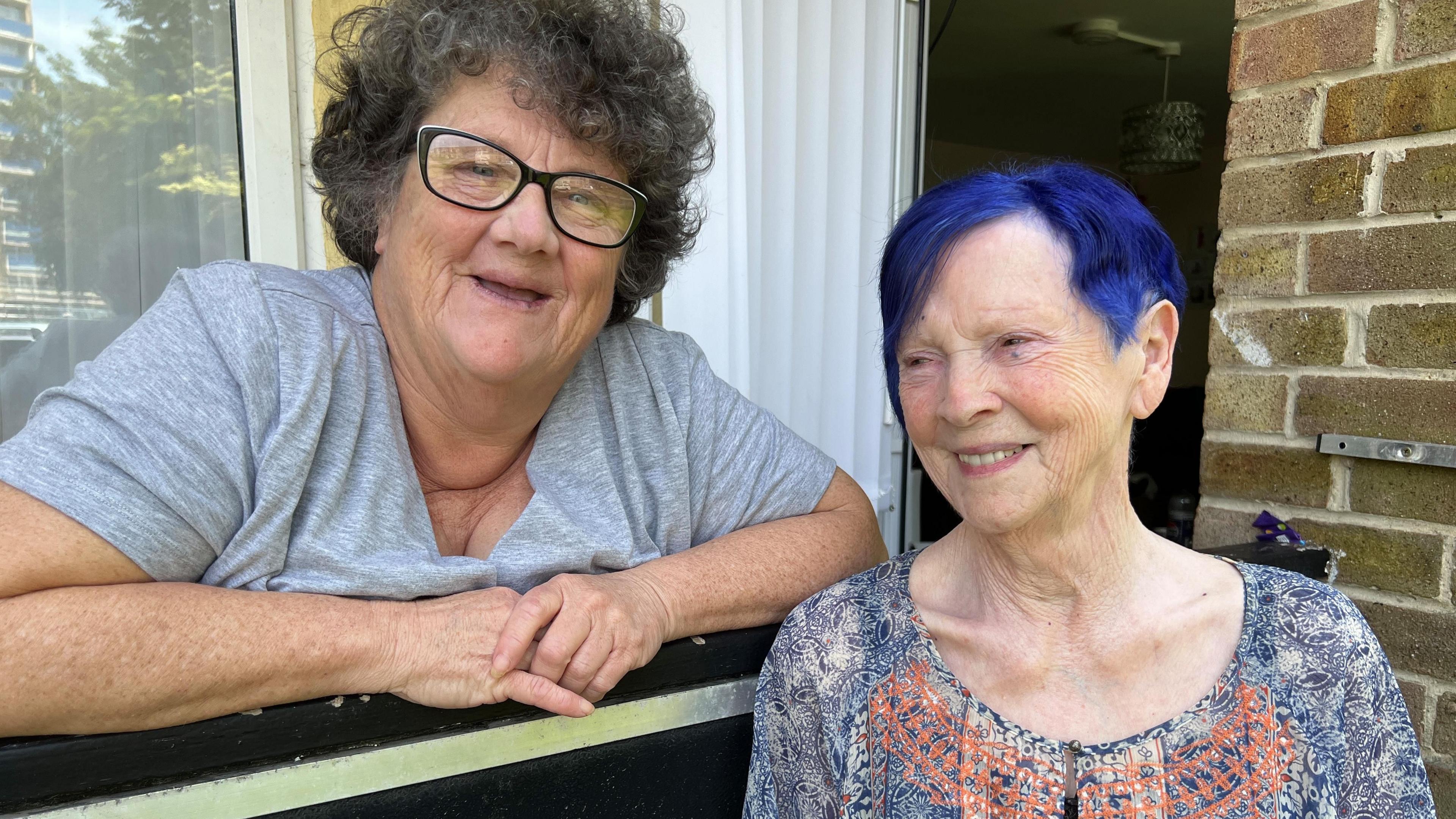 Marie in a grey t-shirt smiles over her first floor balcony alongside Thelma, with bright blue hair on her right.