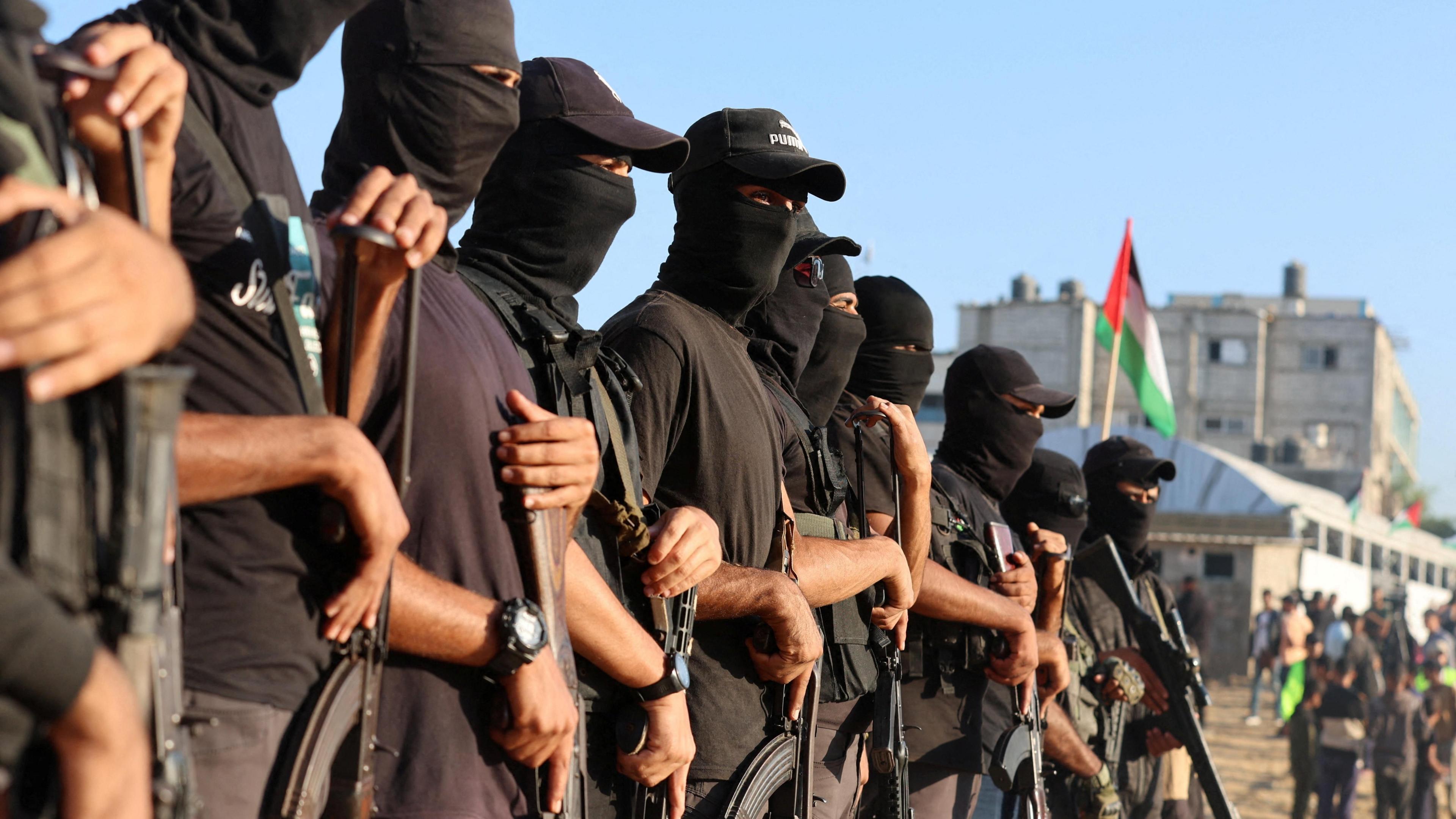 Men wearing black hats and black shirts hold weapons and a Palestinian flag is seen in the background