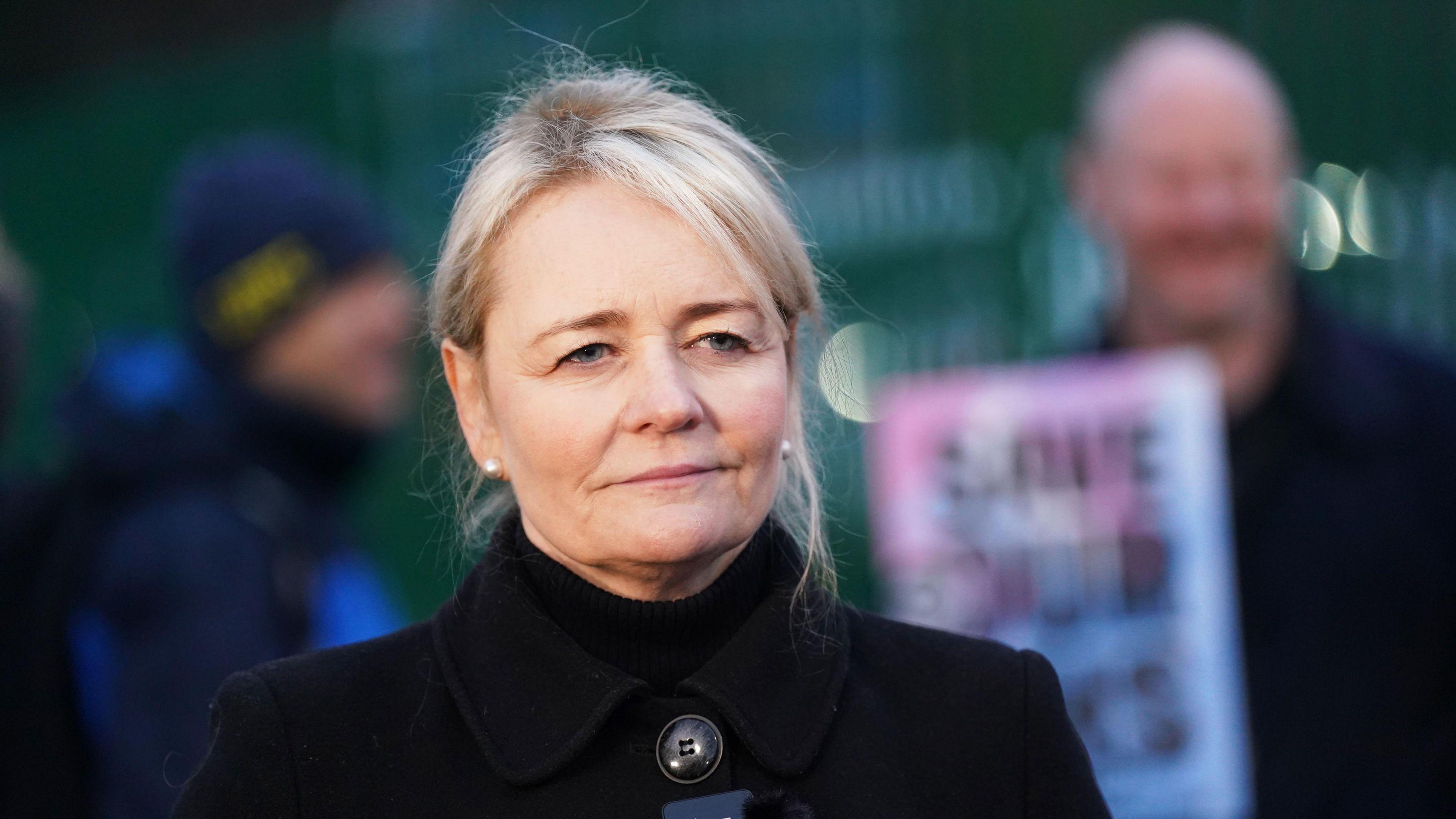 A woman with blonde hair and wearing a black coat, stands in front of a picket line.