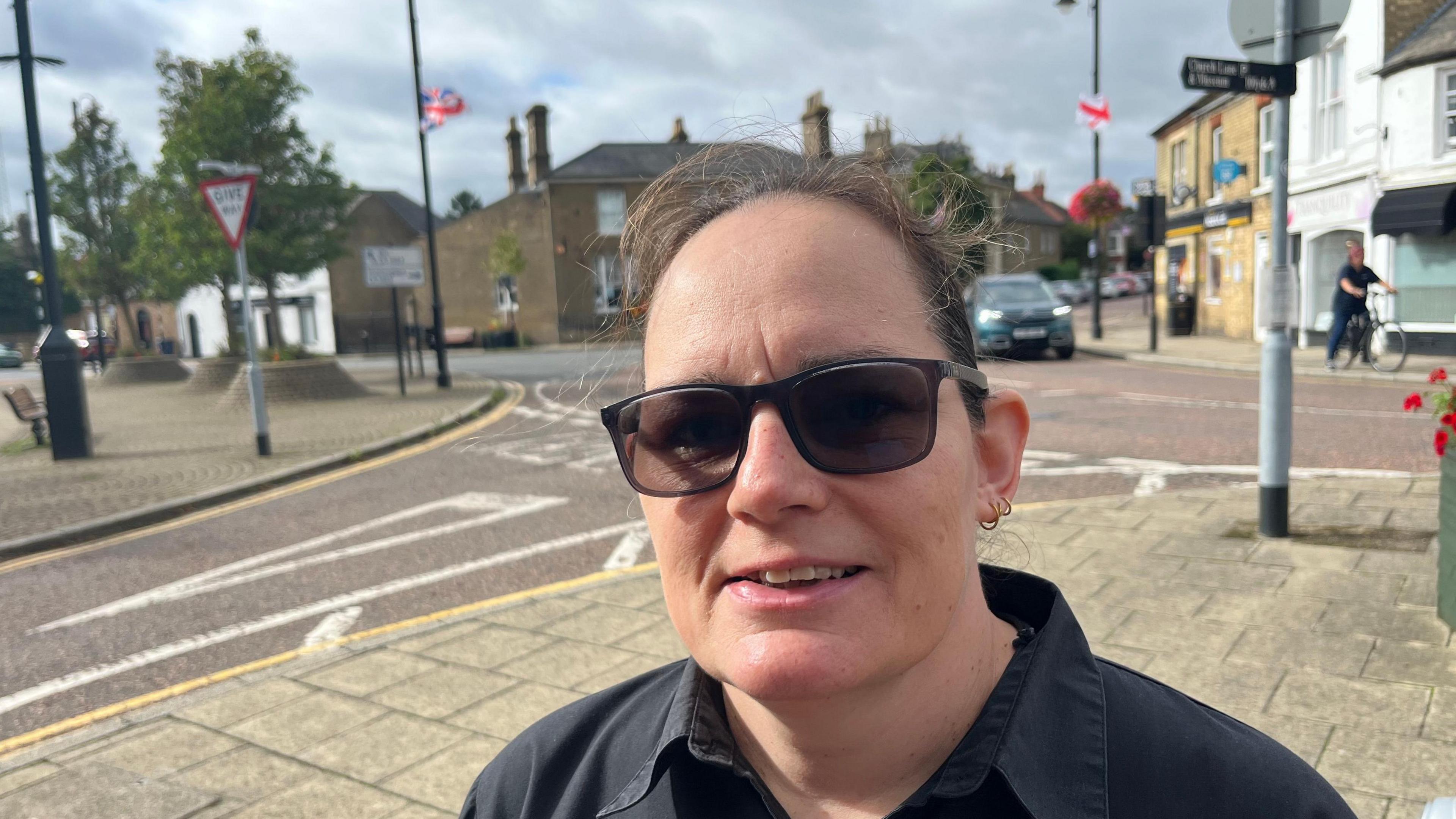 Tina Prior has black short hair swept across her head. She has three small earrings in her left ear lobe. She is wearing dark glasses and a black collared shirt top. Behind her are some union and St George flags hanging from lamp-posts, and there is a road junction visible with shops and houses too.