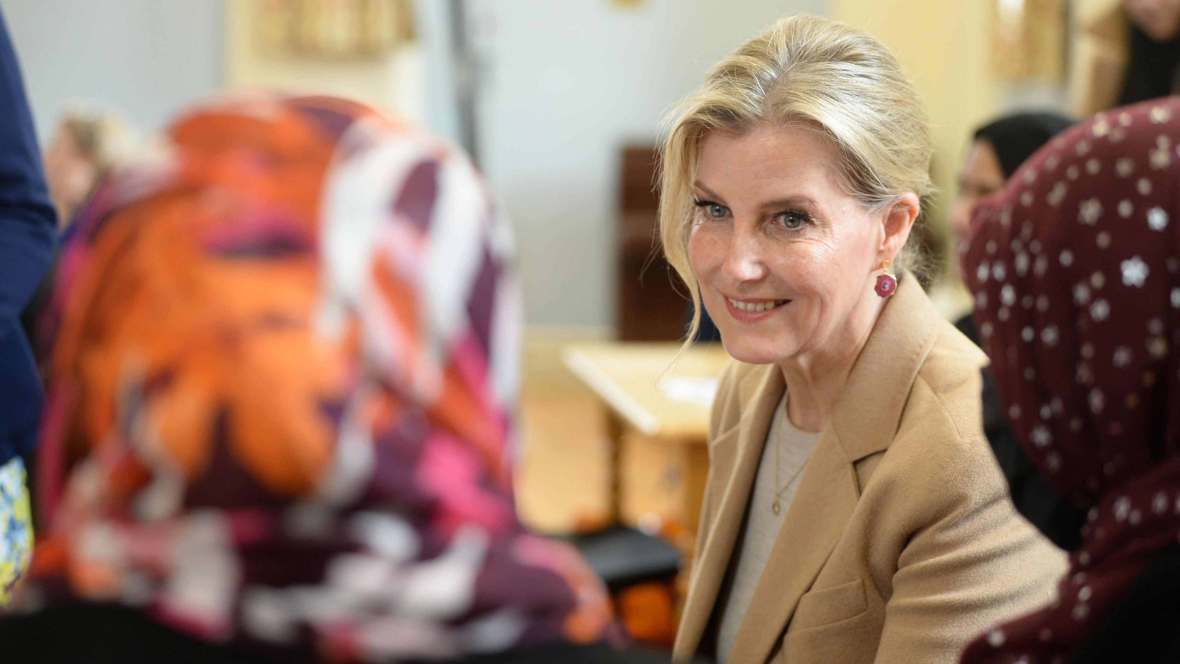 The Duchess of Edingburgh visiting the English Language club in April. Her blonde hair is pulled pack with a tendril at the front of her face, She is wearing a light coloured knit with a camel coat and dainty gold pendant