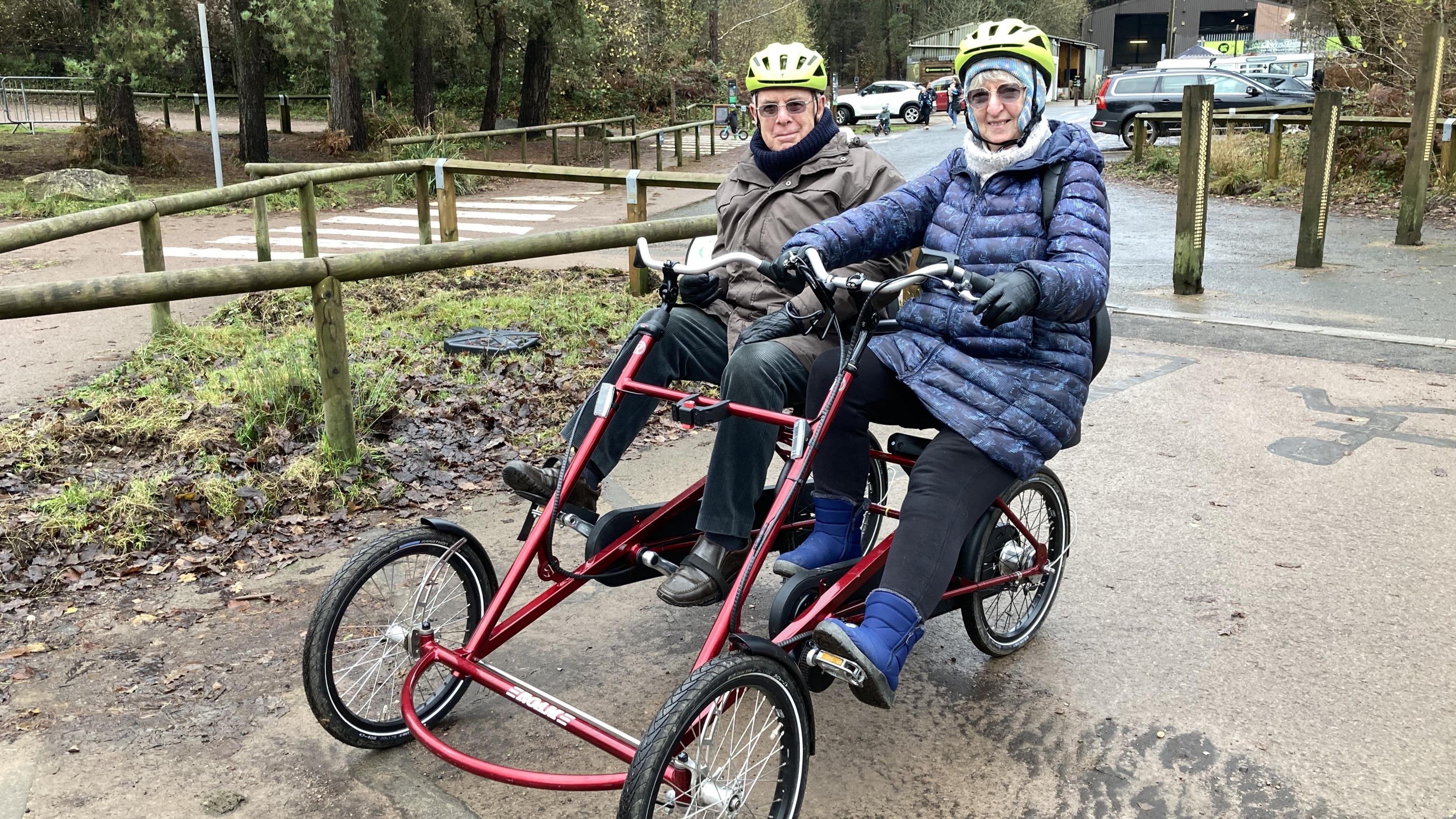 Kathryn and Roger Bellchamber sitting next to each other on a side by side red bike. They are both wearing yellow helmets and they have dark focal lens glasses. Kathryn has a light blue hat under her helmet and is wearing dark blue boots and a blue patterned coat. They are both smiling at the camera.