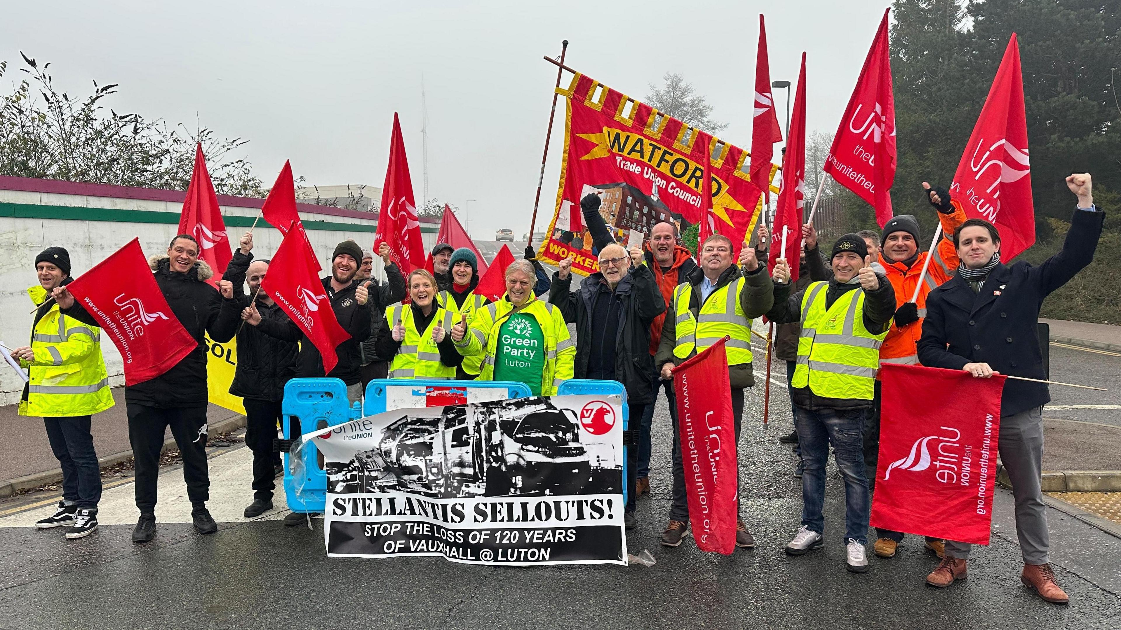 A large group of protesters with red flags stand together outside the Vauxhall plant in Luton