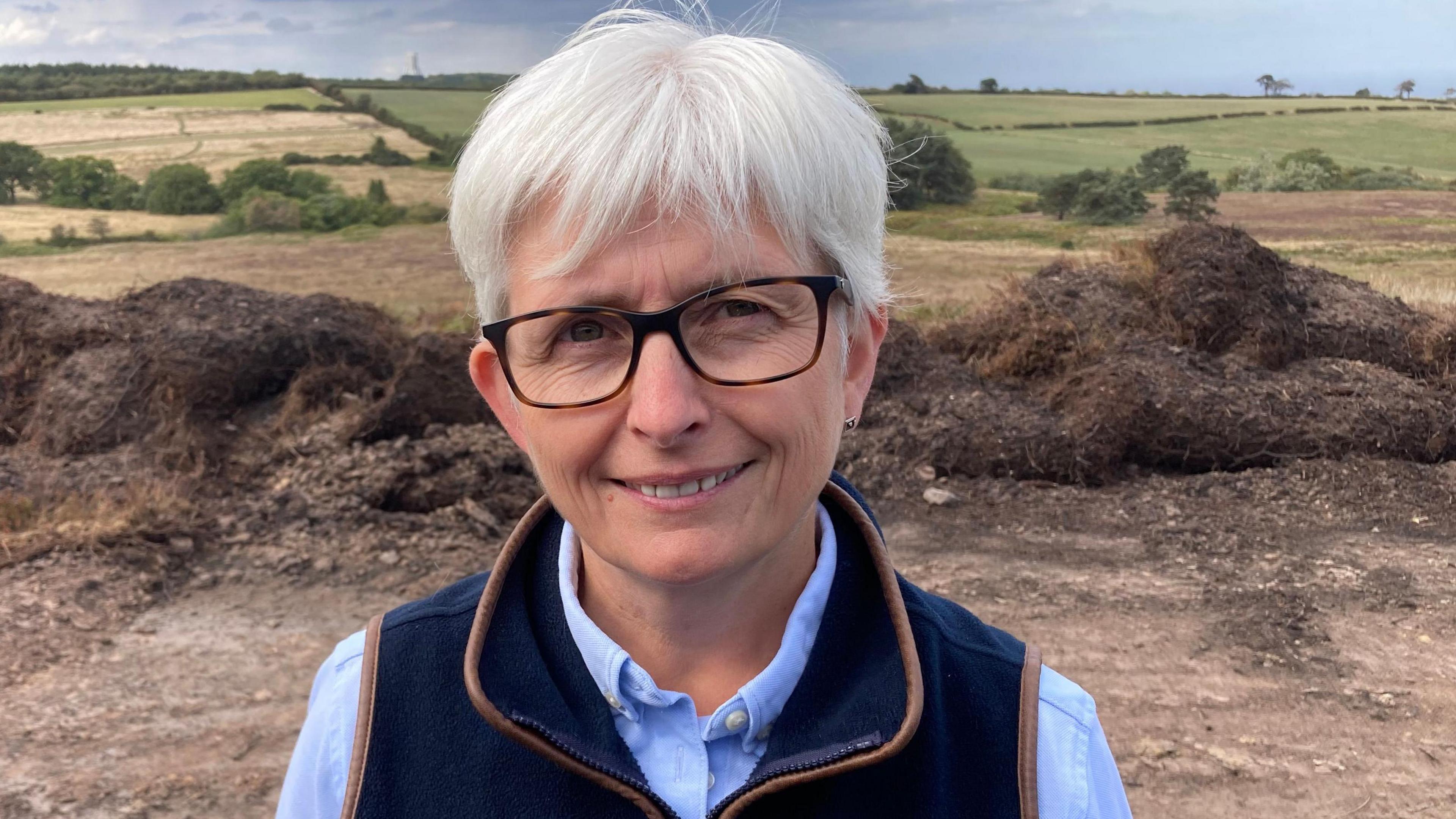 A woman with grey hear and wearing glasses smiles at the camera, with moorland in the background.