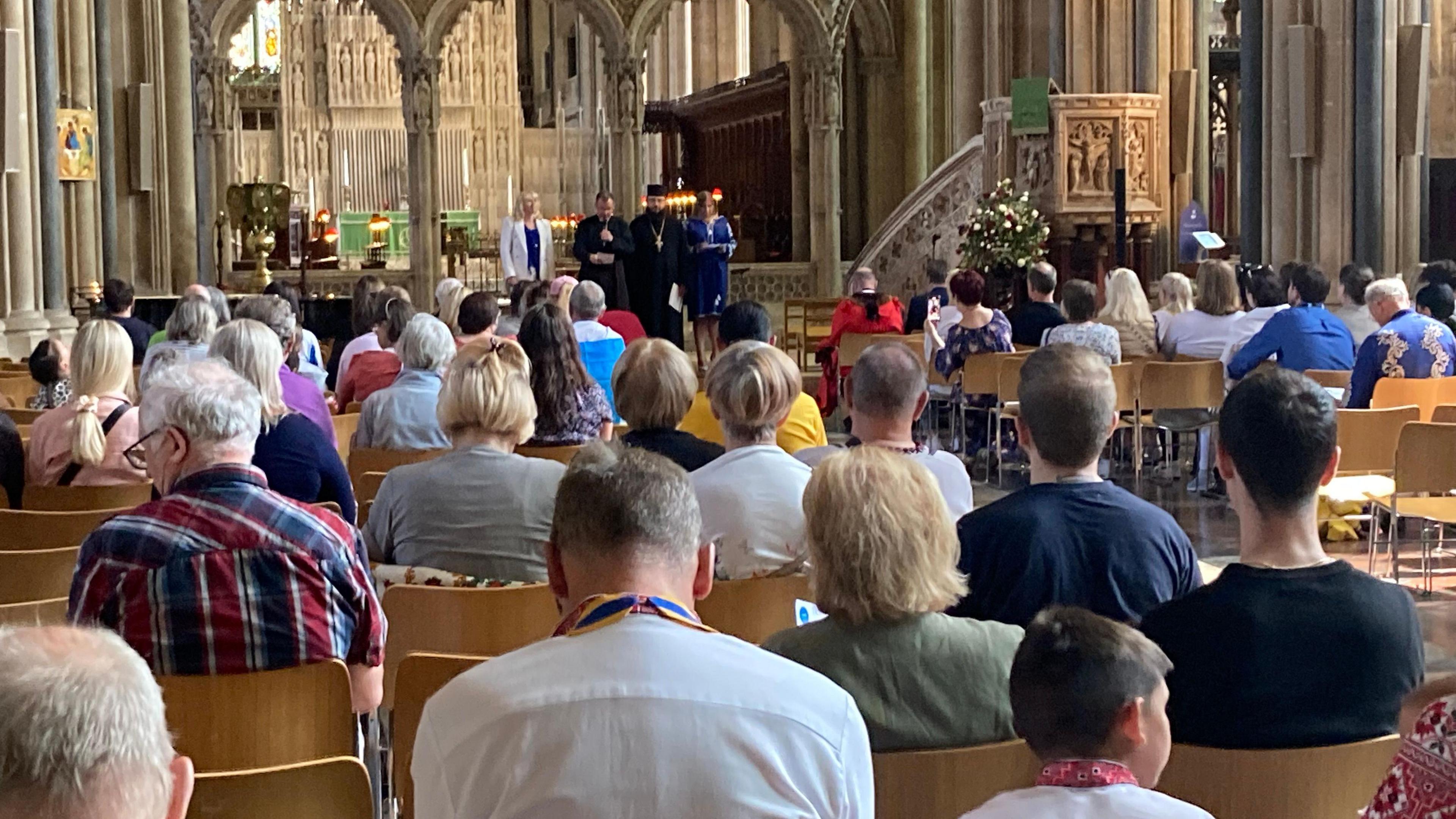 An audience is seen from behind, sitting on wooden chairs inside Bristol Cathedral. They are watching four people standing at the cathedral's altar. Lots of stone pillars are in the background.