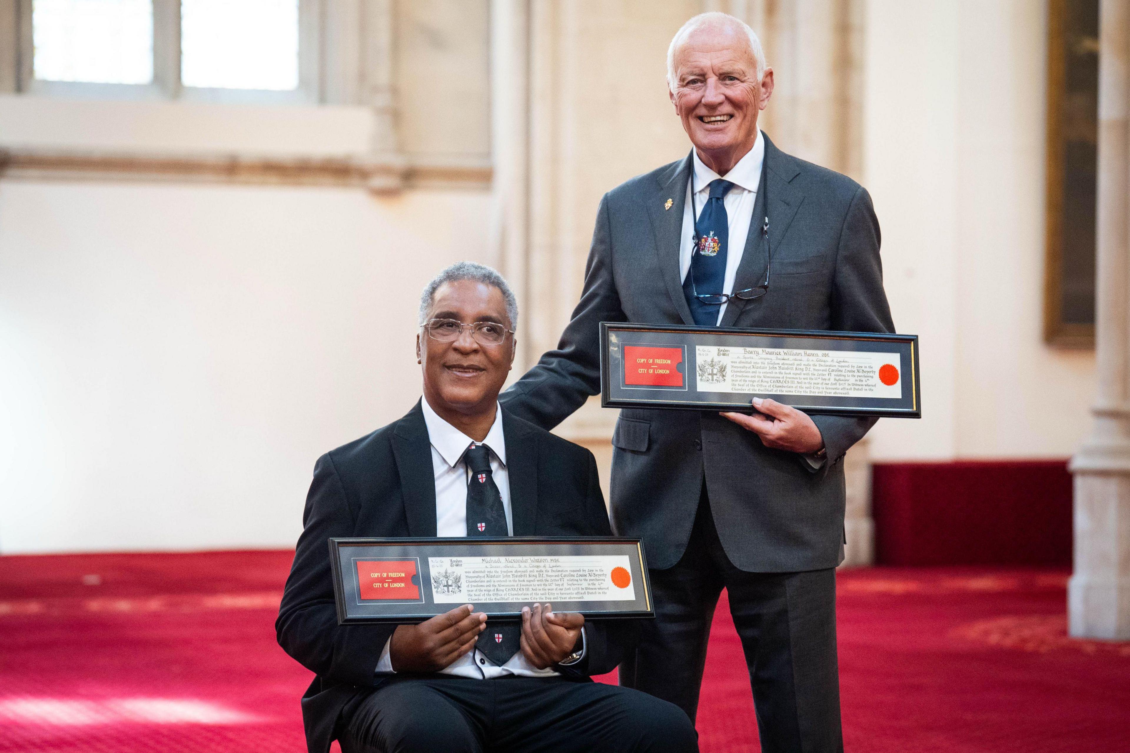 Michael Watson and Barry Hearn who are both wearing suits stand in the Guildhall holding the framed documents of their Freedom to the City. Michael Watson is sitting in a wheelchair while Barry Hearn is standing beside him and has his hand on Michael's back