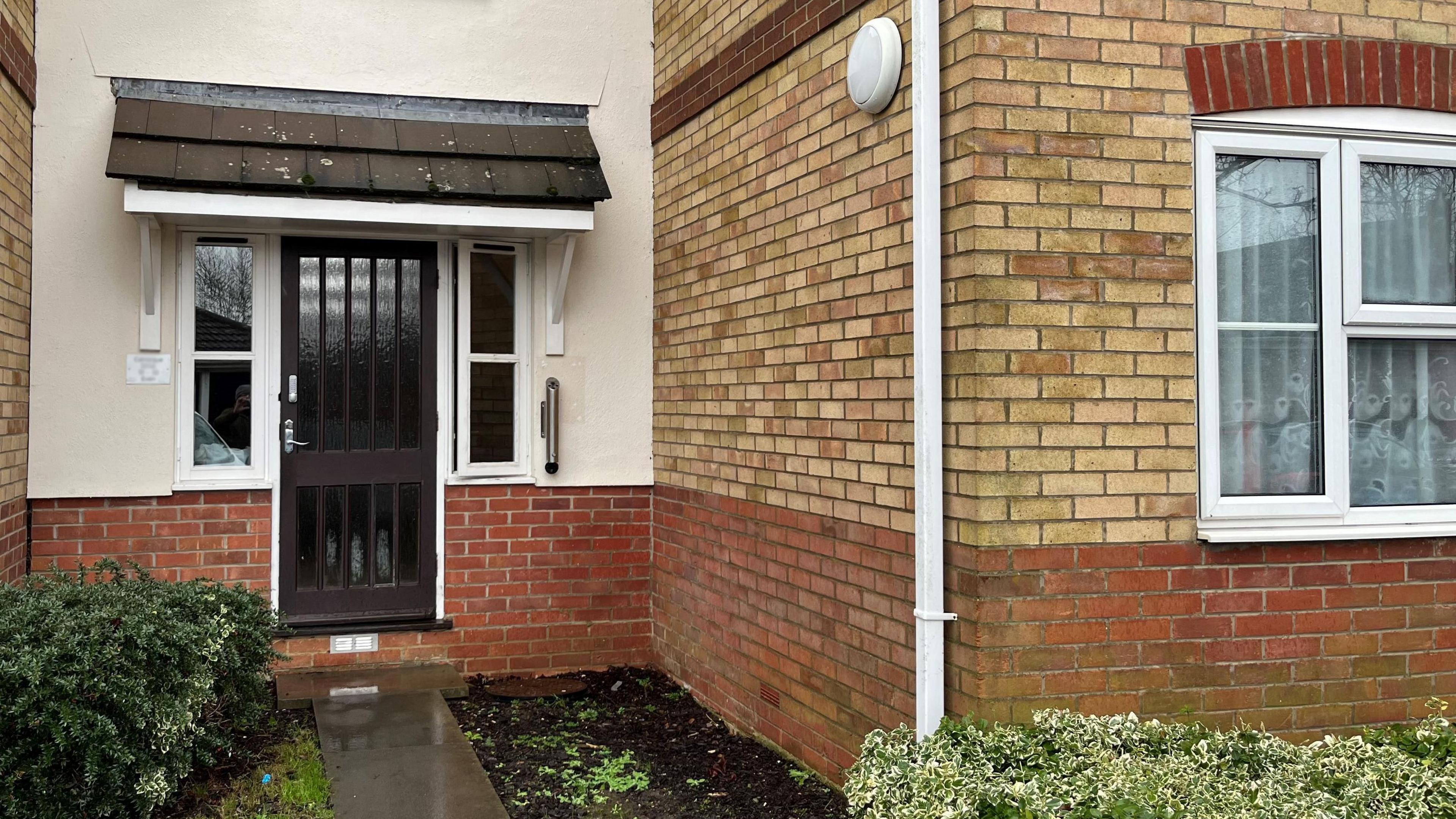 The outside of a ground floor flat, with hedges and shrubs growing at the front of it. There is a brown communal front door and the Chapmans live in a one-bedroom ground-floor flat on the right.