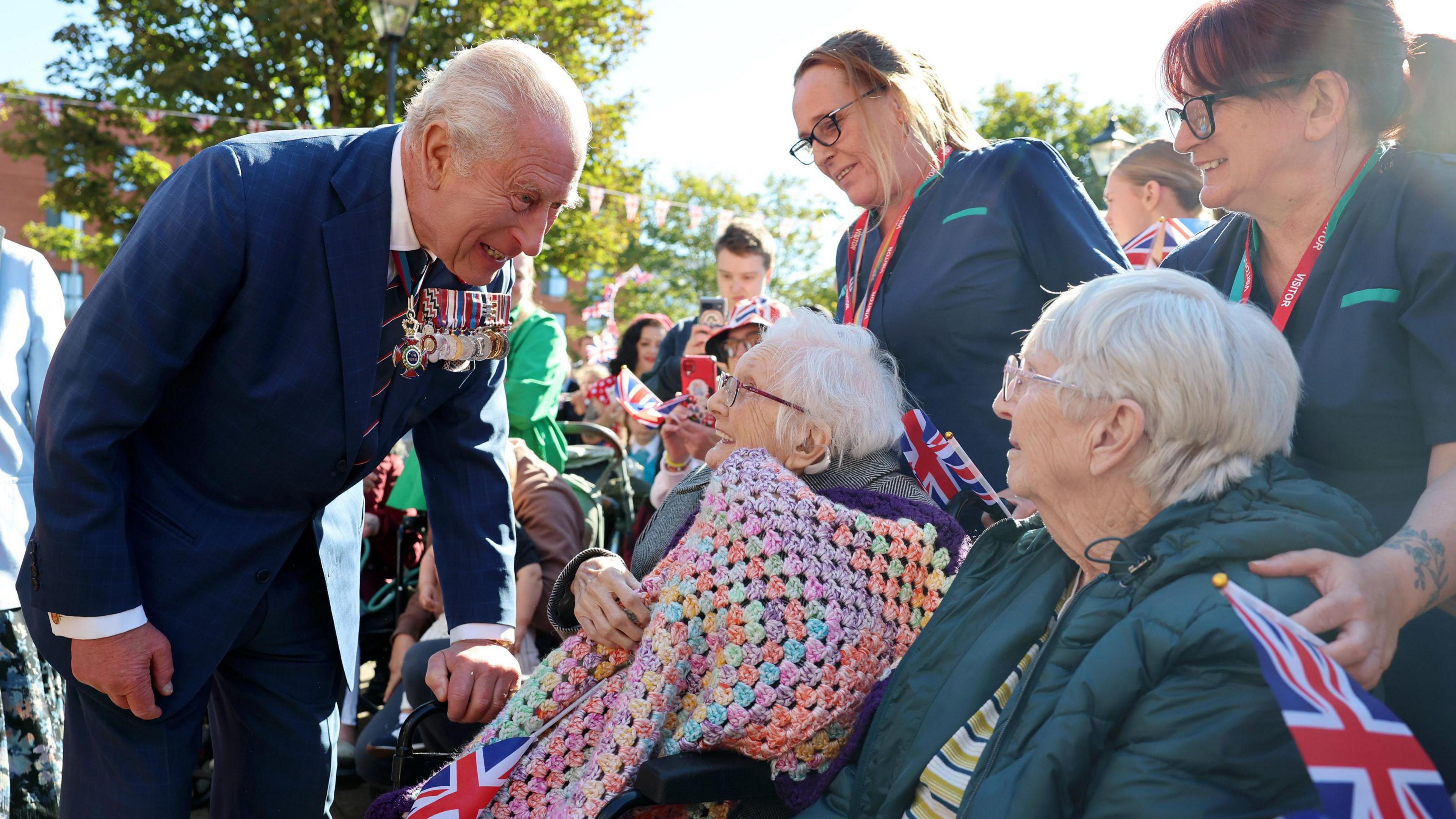King Charles III meets members of the public during a visit to Barrow-in-Furness. The King, who has a huge row of medals on this jacket, is leaning down to speak to a lady in a wheelchair, who is smiling and holding a colourful knitted blanket. There are lots of people milling around and lots of union jacks.